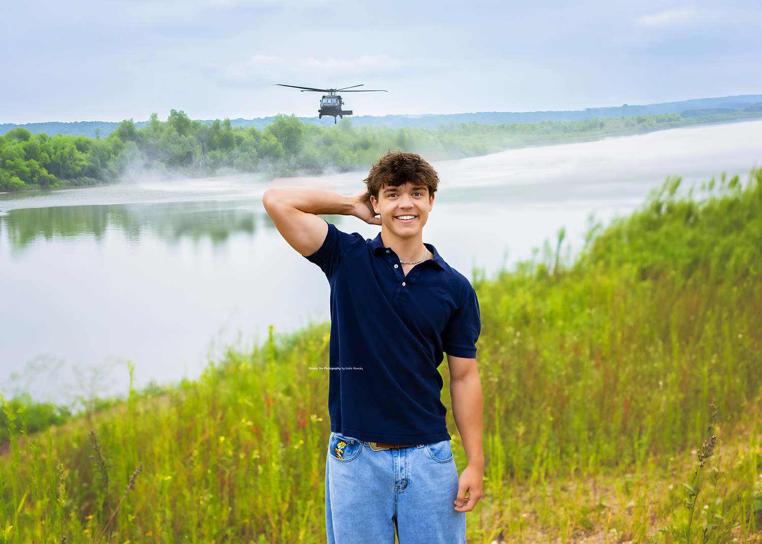 Young man smiling outdoors near a river with a helicopter flying in the background on a cloudy day.
