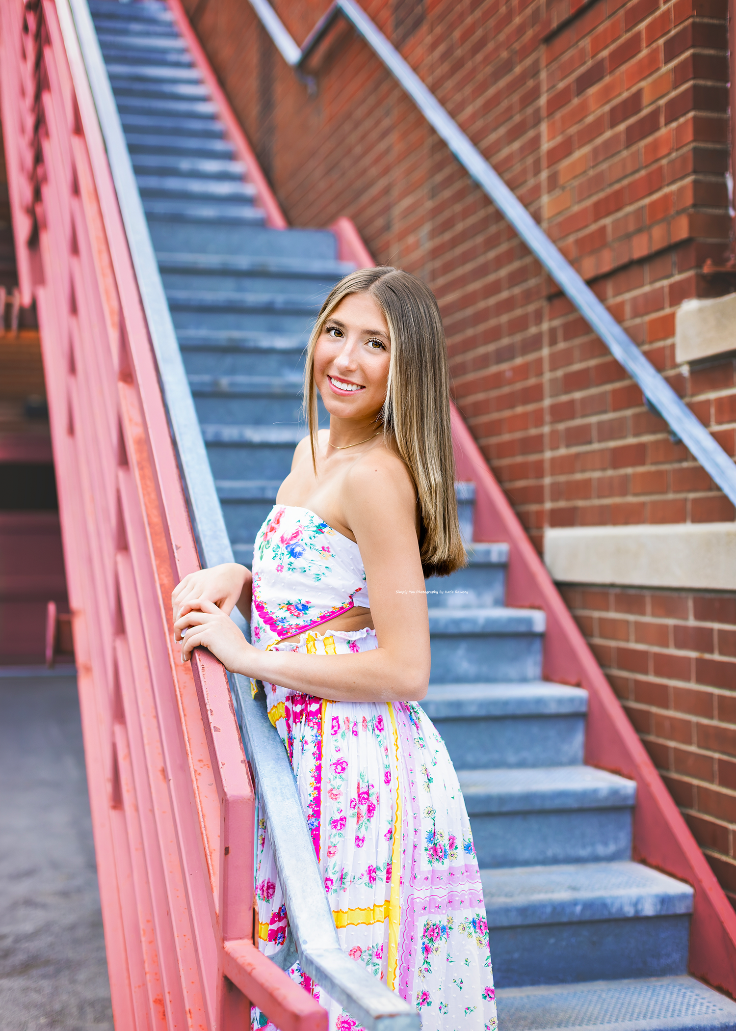 Smiling young woman in floral dress posing on pink metal staircase against brick wall.