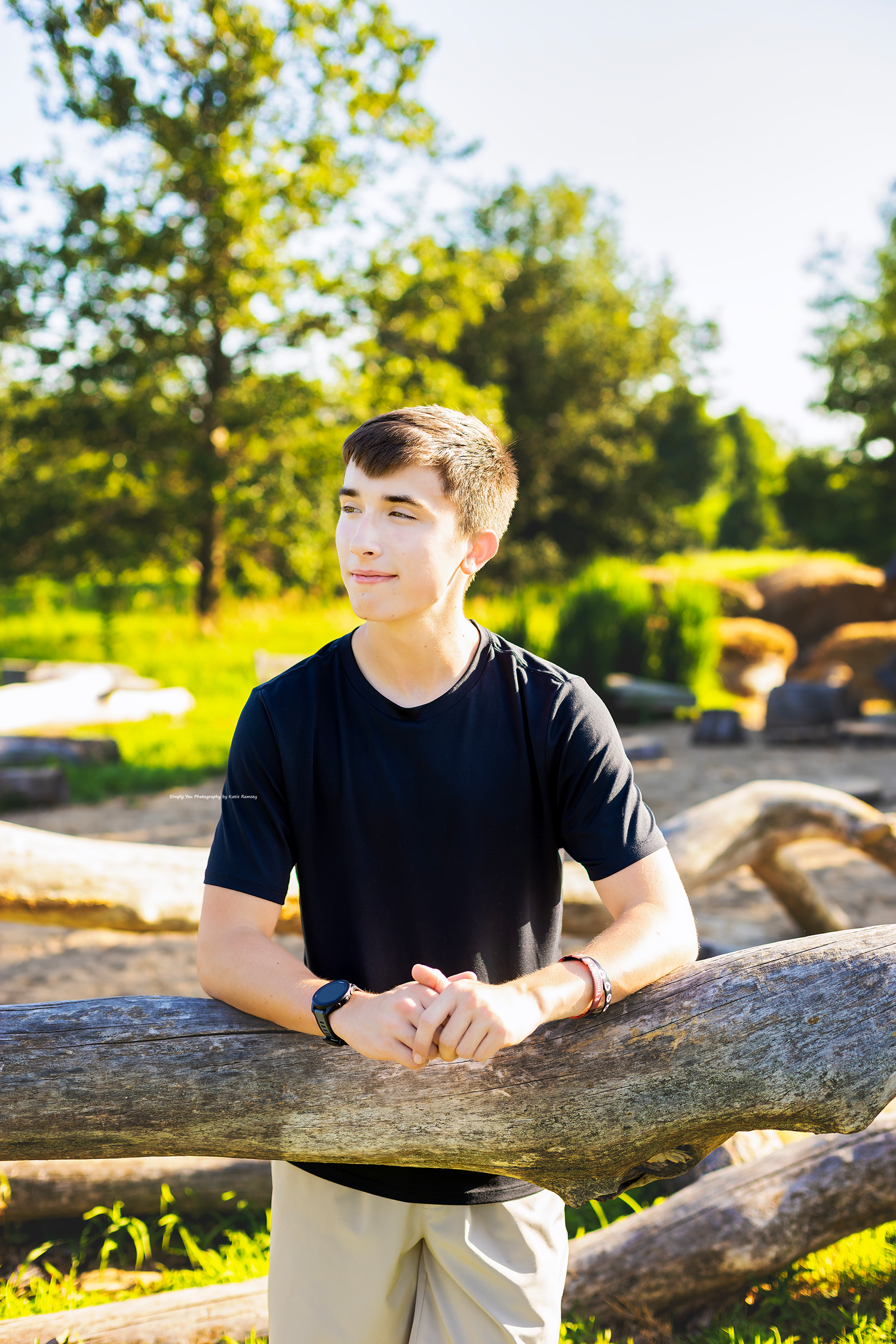 A teenage boy in a black t-shirt leans on a wooden log fence in a sunny park setting.