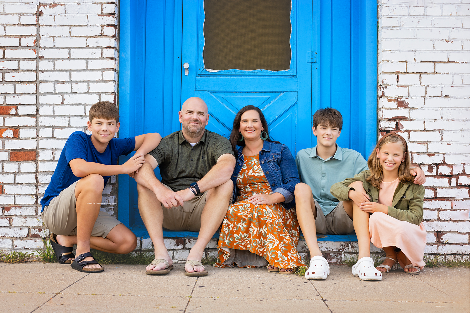Family of five smiling together in front of a blue door on a brick wall outdoors.