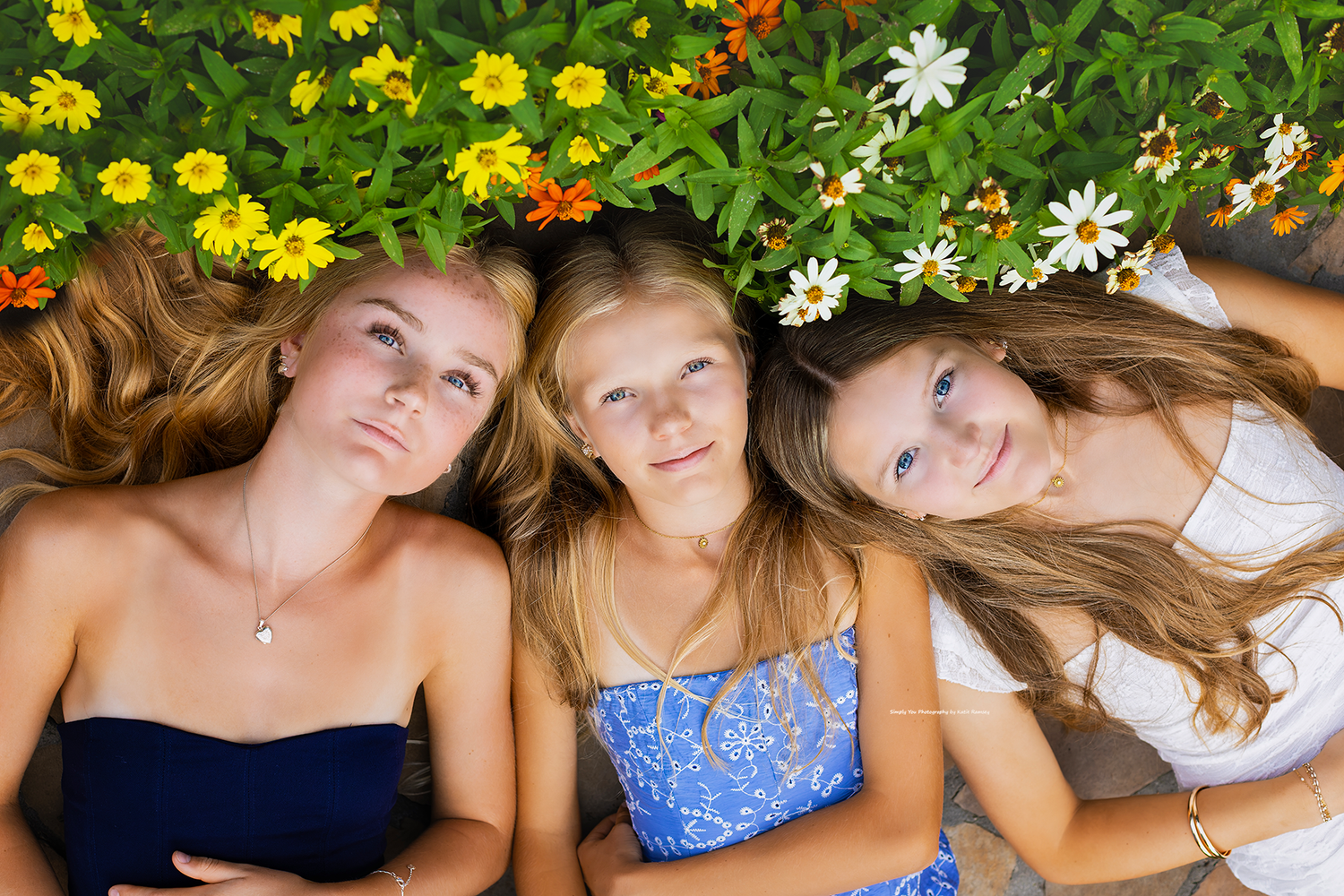 Three teenage girls lying on grass surrounded by colorful yellow and white flowers, smiling up at the camera.