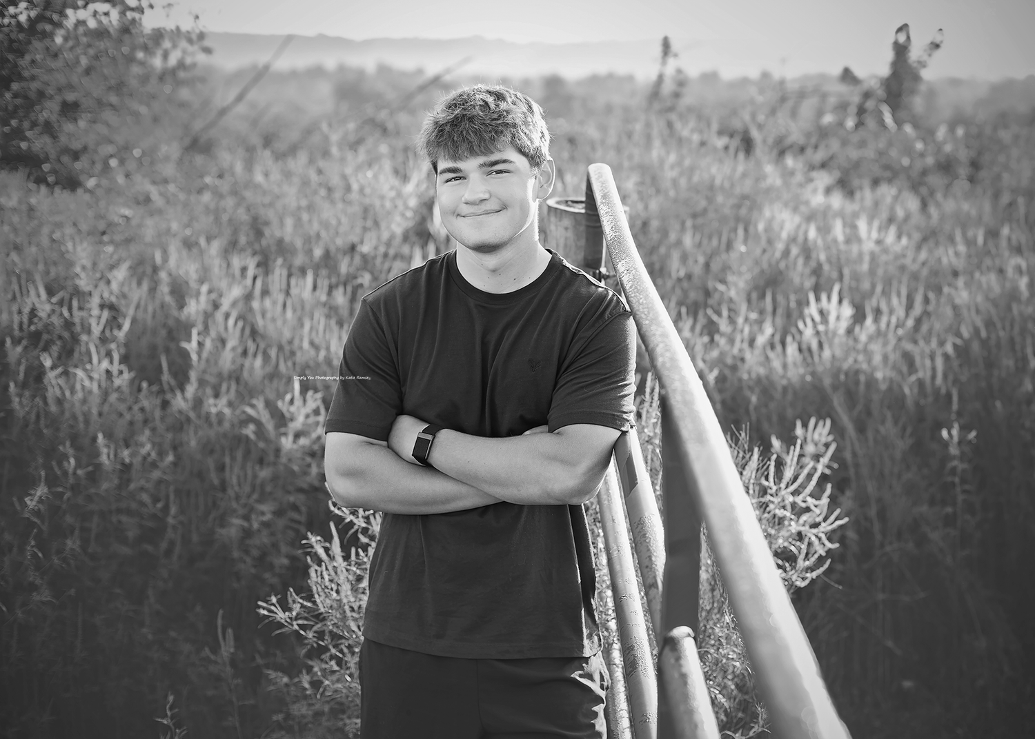 Black and white photo of a smiling young man with crossed arms standing in a field near a wooden fence.