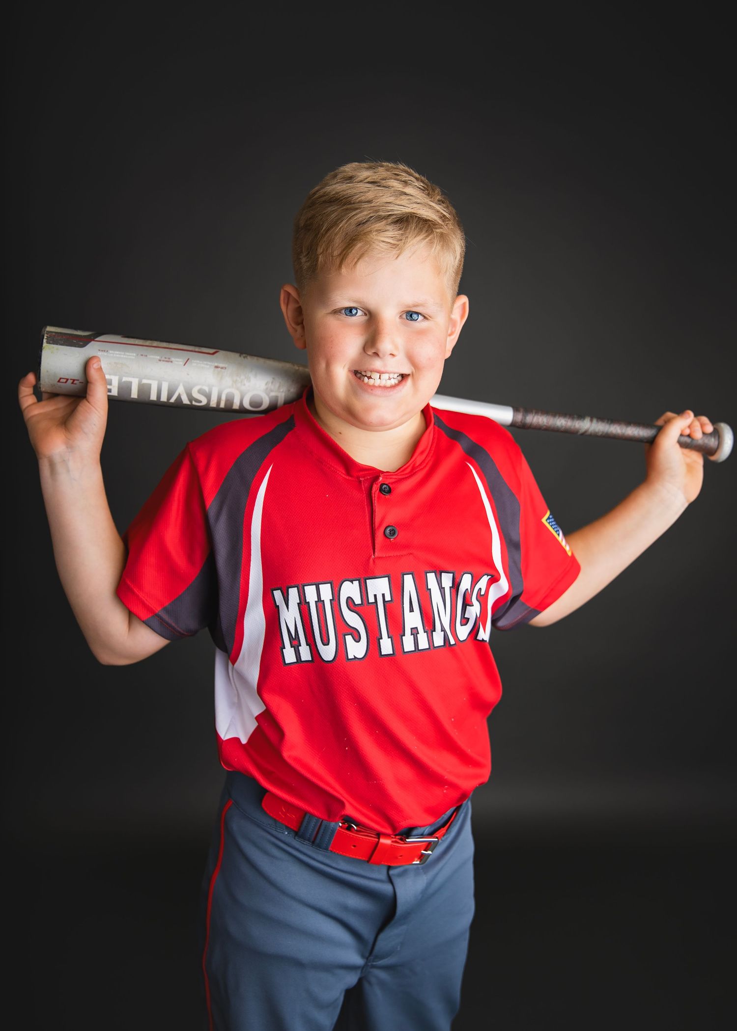 Smiling young boy in red Mustangs baseball jersey poses with Louisville Slugger bat across shoulders.