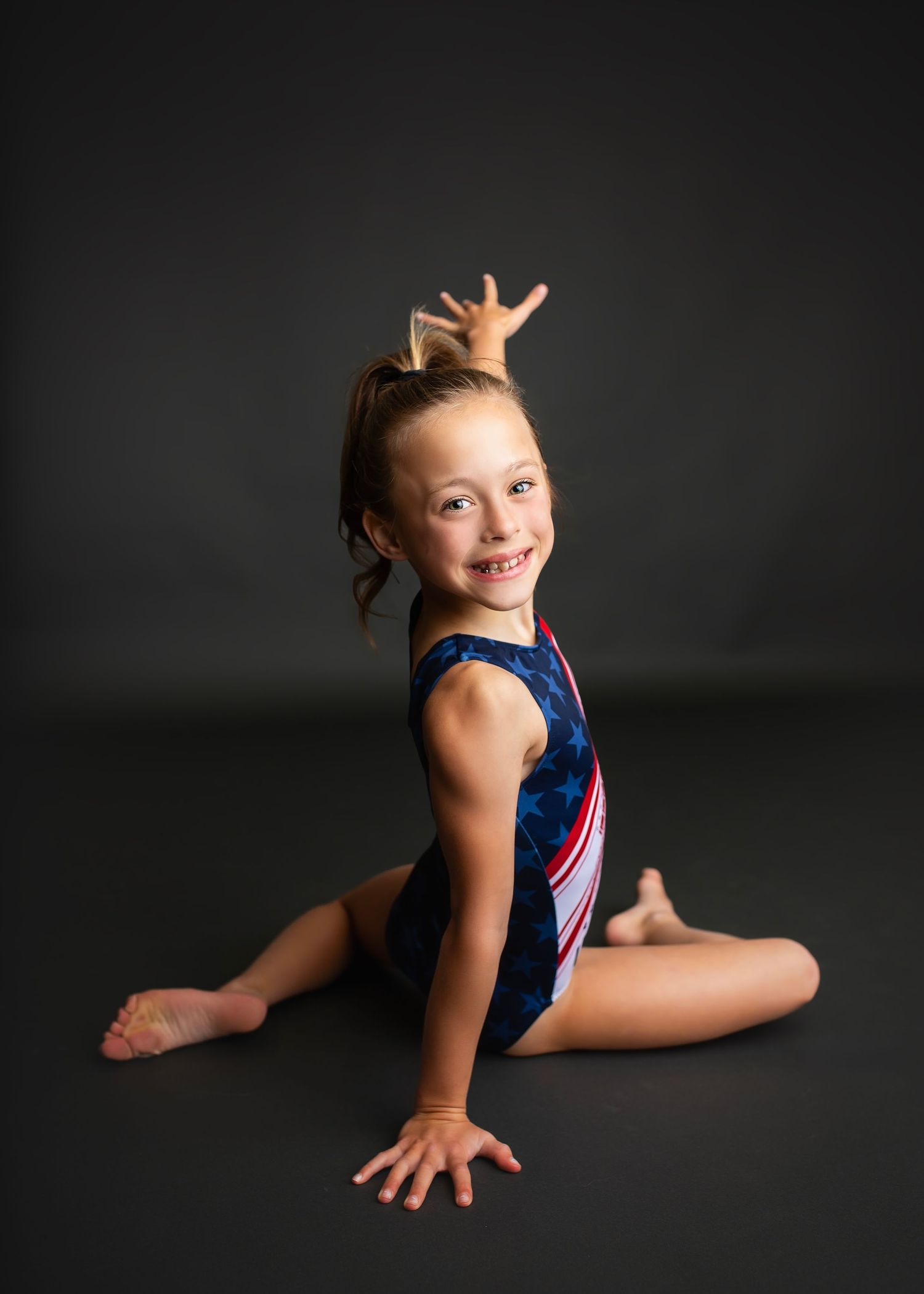 Young gymnast in USA leotard sitting on floor smiling with legs crossed against dark background.