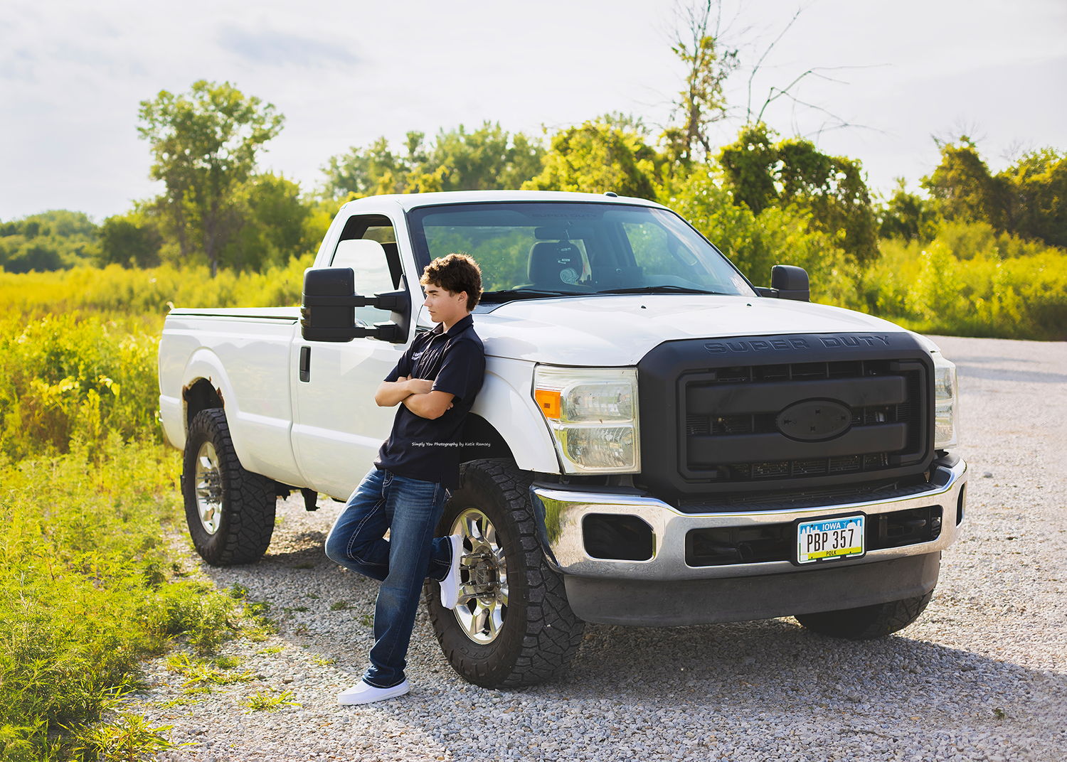 Young person leaning against a white Ford F-250 pickup truck parked on a gravel road surrounded by green trees.