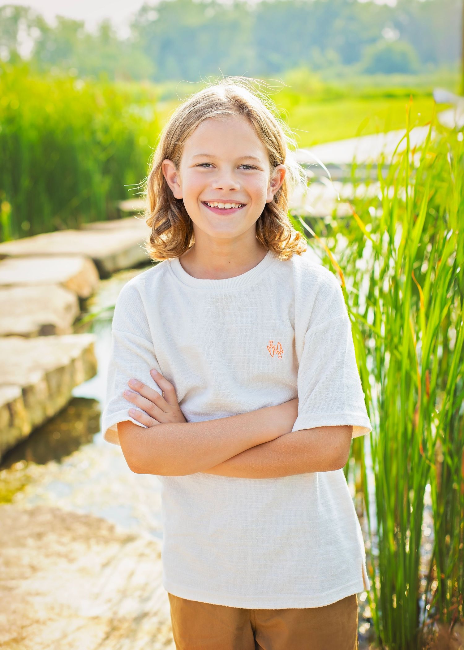 Smiling boy with long hair standing arms crossed near a pond with tall green reeds in sunlight.