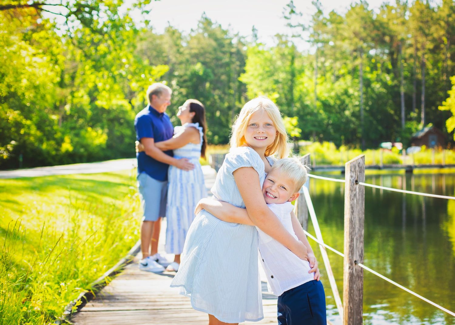 Two blonde siblings hug on a sunny dock while their parents embrace in the background near a lake.