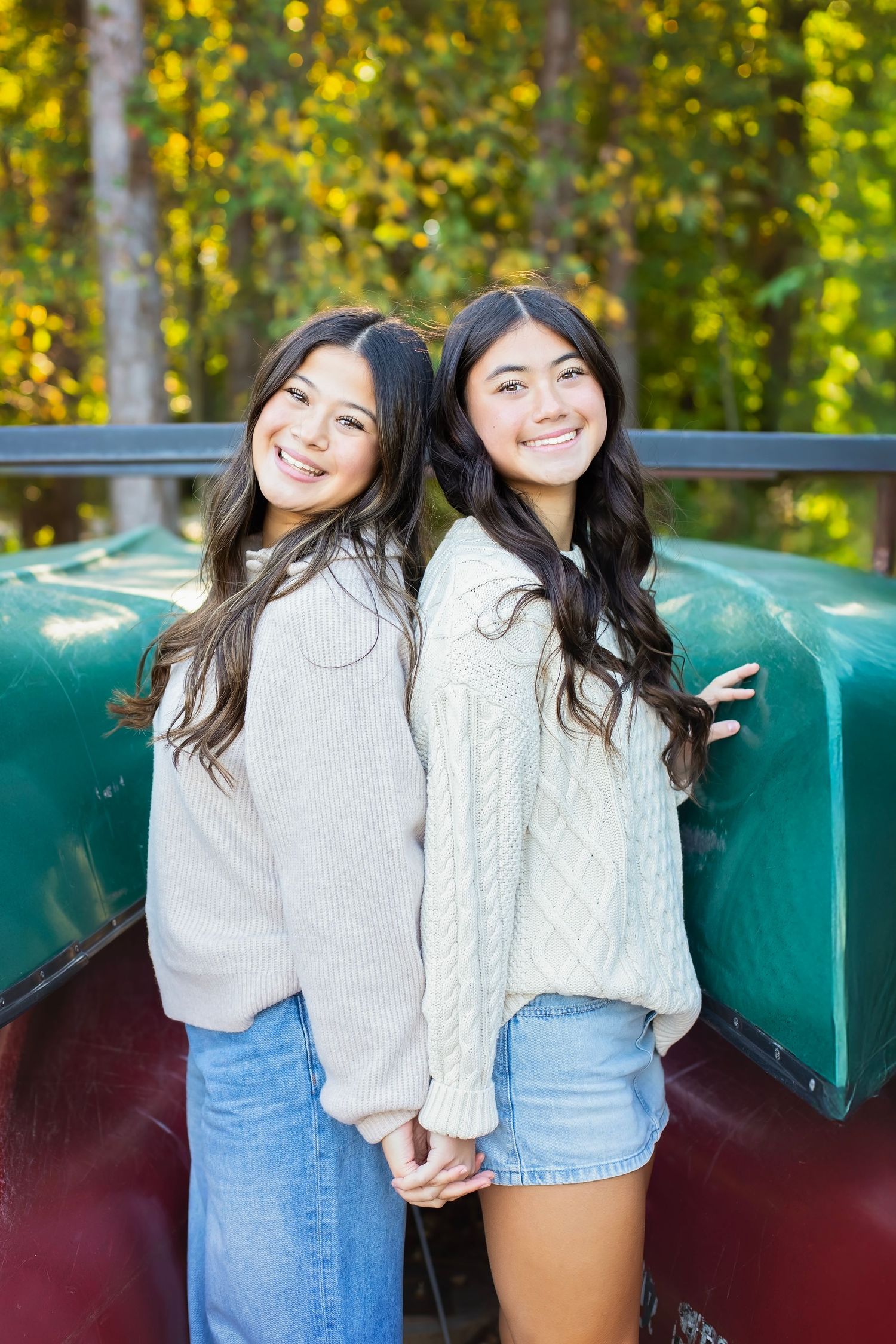 Two teenage girls in cozy sweaters smiling back-to-back near green canoes outdoors.