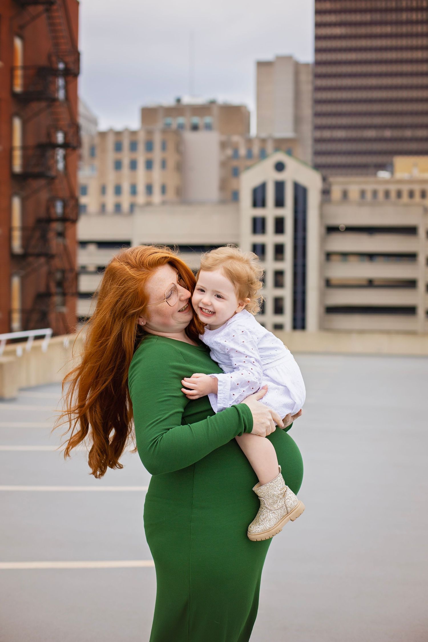 Pregnant redhead mother in green dress holding laughing toddler on urban rooftop with city skyline.
