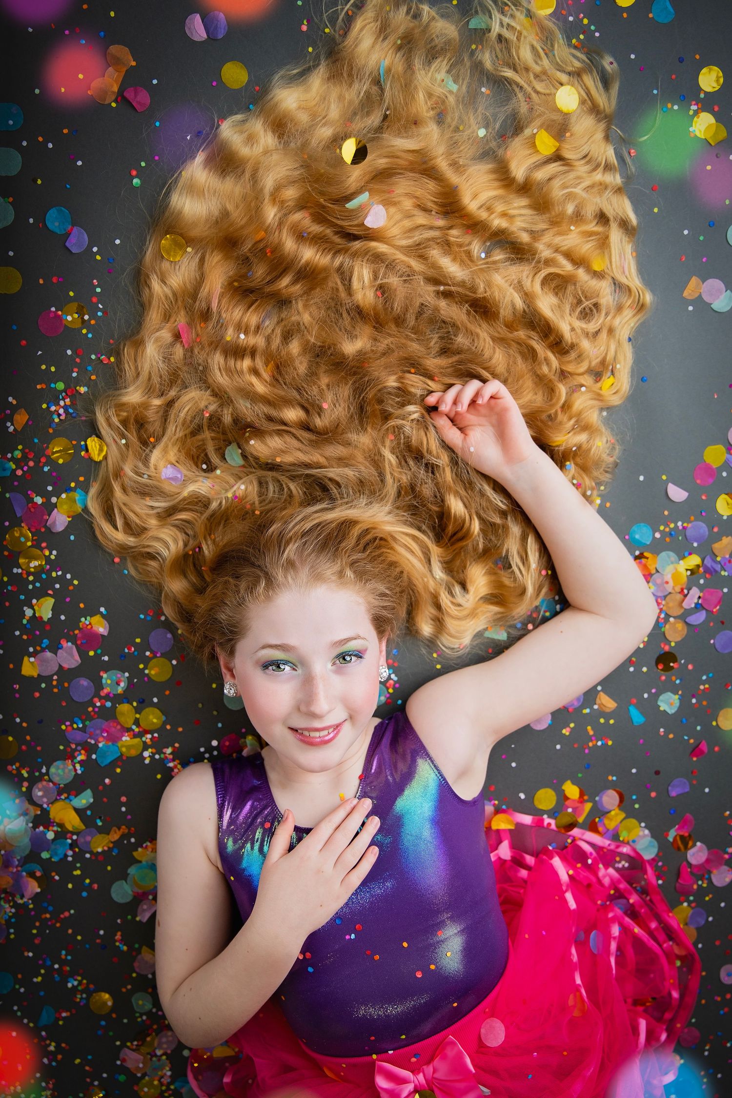 A young girl with voluminous curly hair lies on a dark floor in a purple top and pink skirt with confetti.