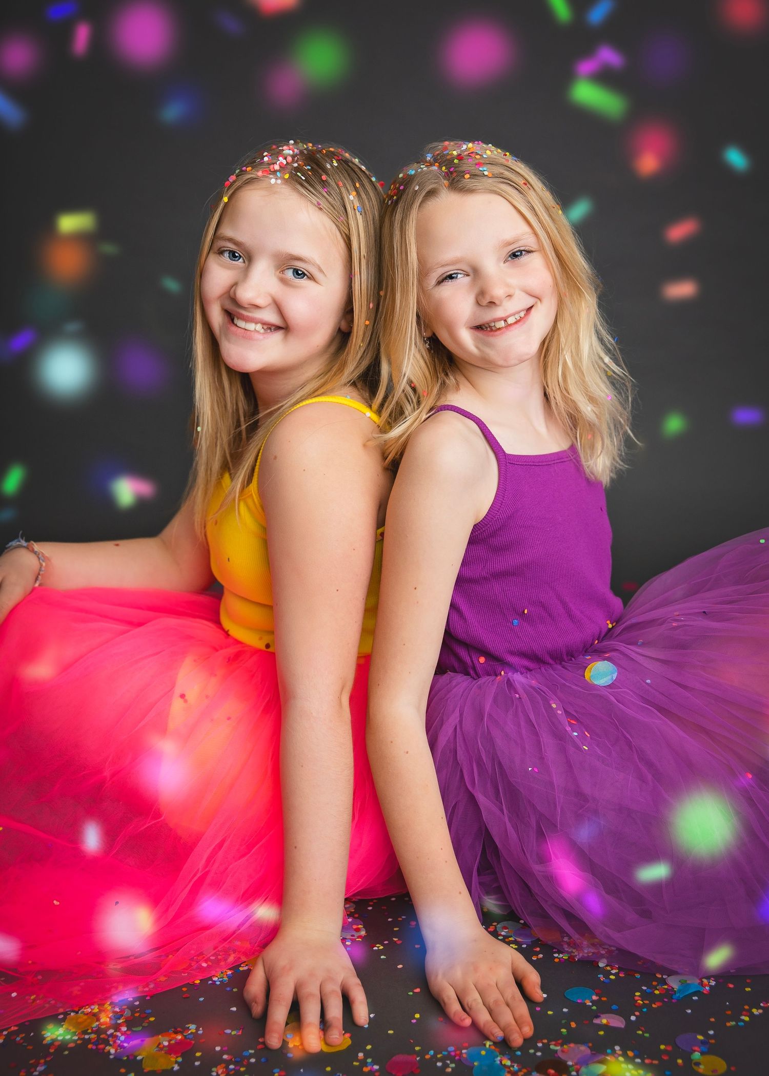 Two blonde sisters in bright tutus sitting back-to-back smiling with confetti falling around them.