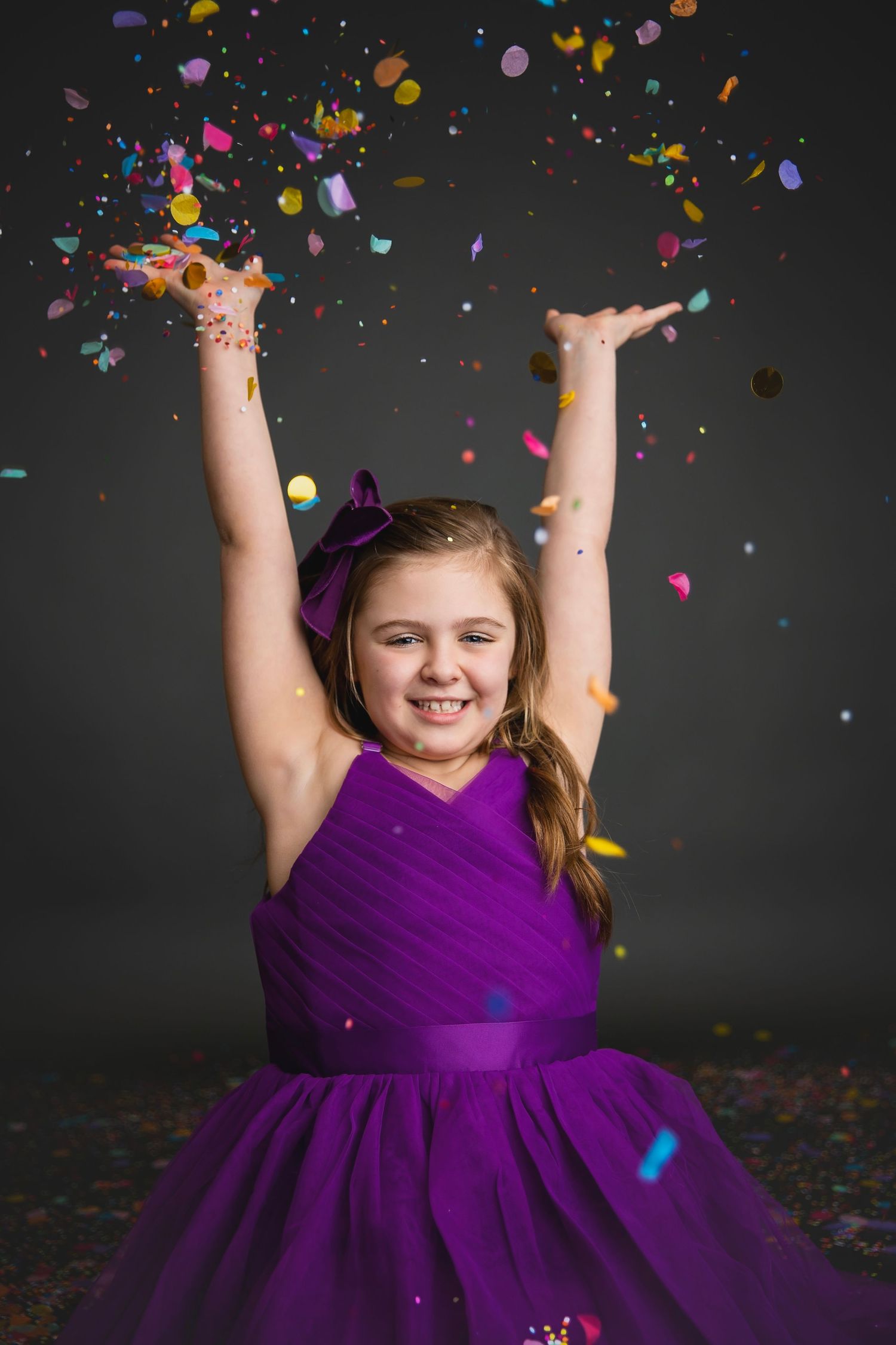Excited girl in purple dress tossing colorful confetti in the air during a festive studio shoot.