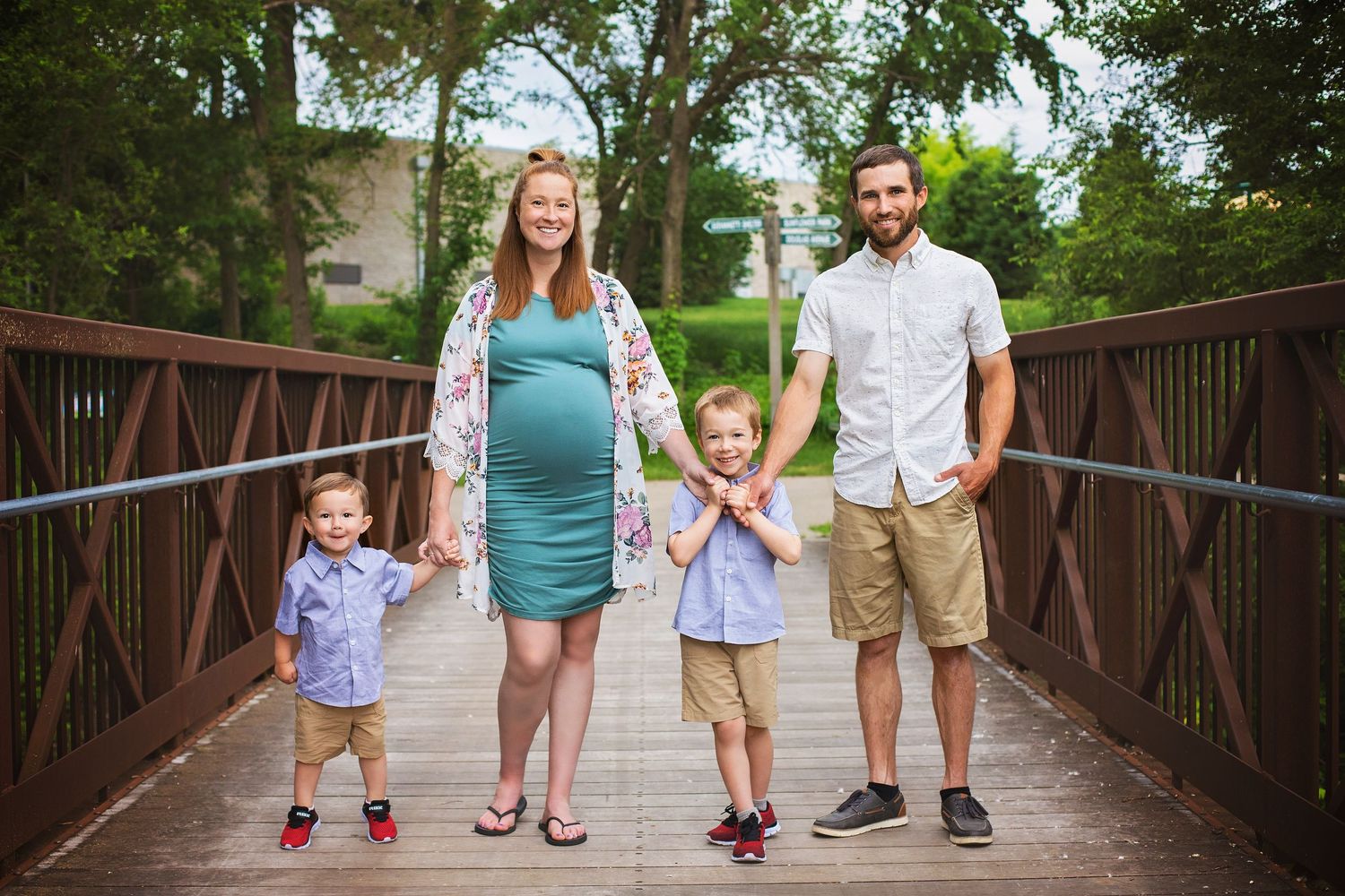Family of four poses on a wooden bridge, with pregnant mother in teal dress surrounded by husband and two young sons.