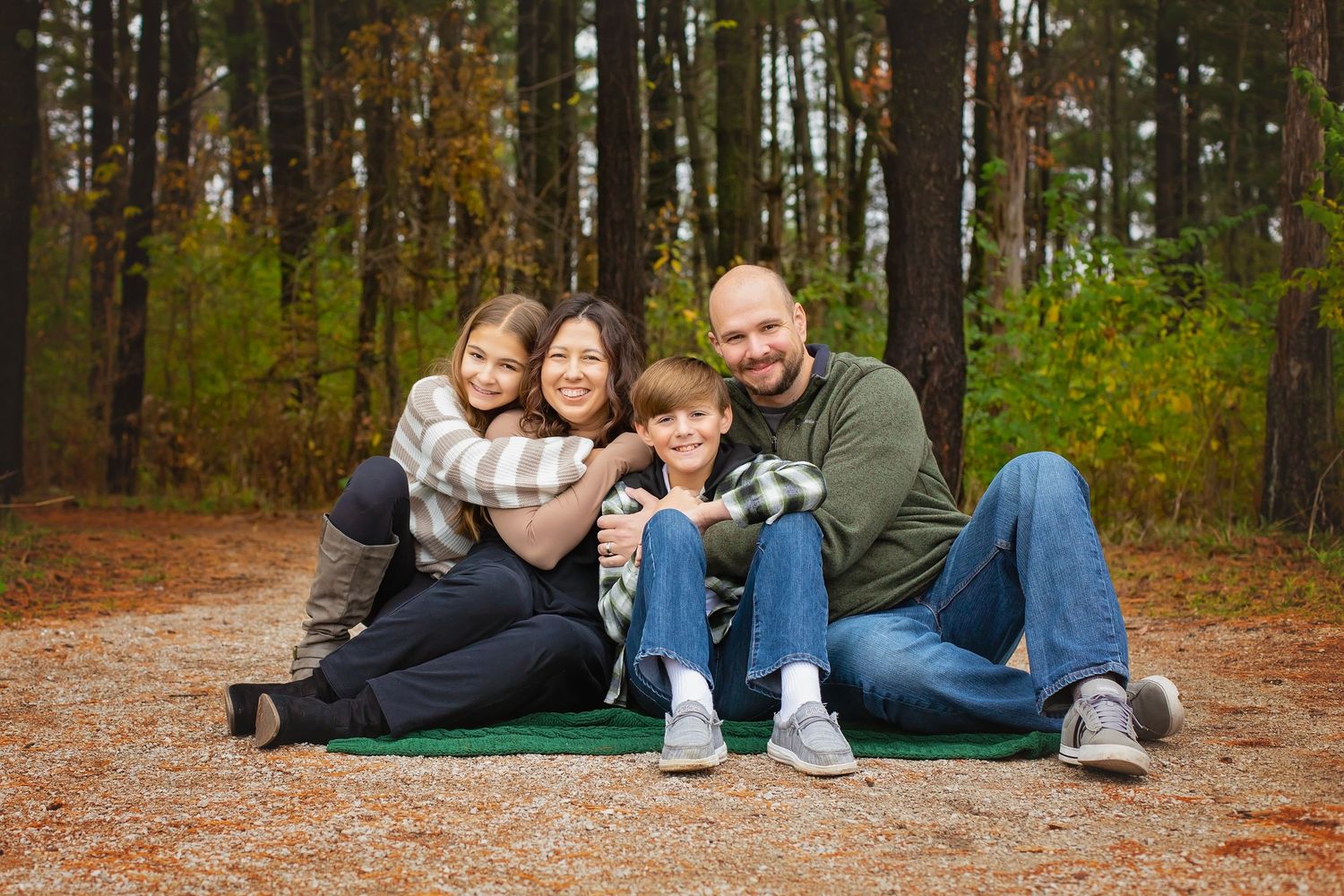 A family of four sits together on a blanket on a forest path surrounded by colorful fall foliage.