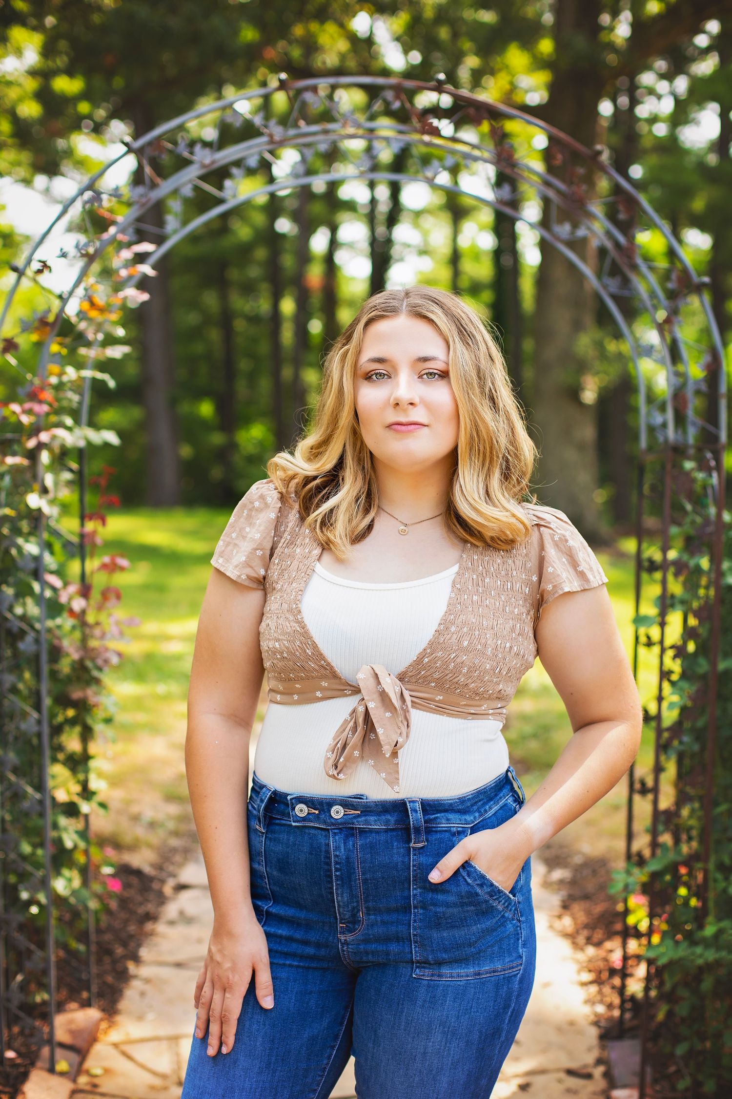 Young blonde woman in crop top and jeans standing under decorative garden arch with flowers.