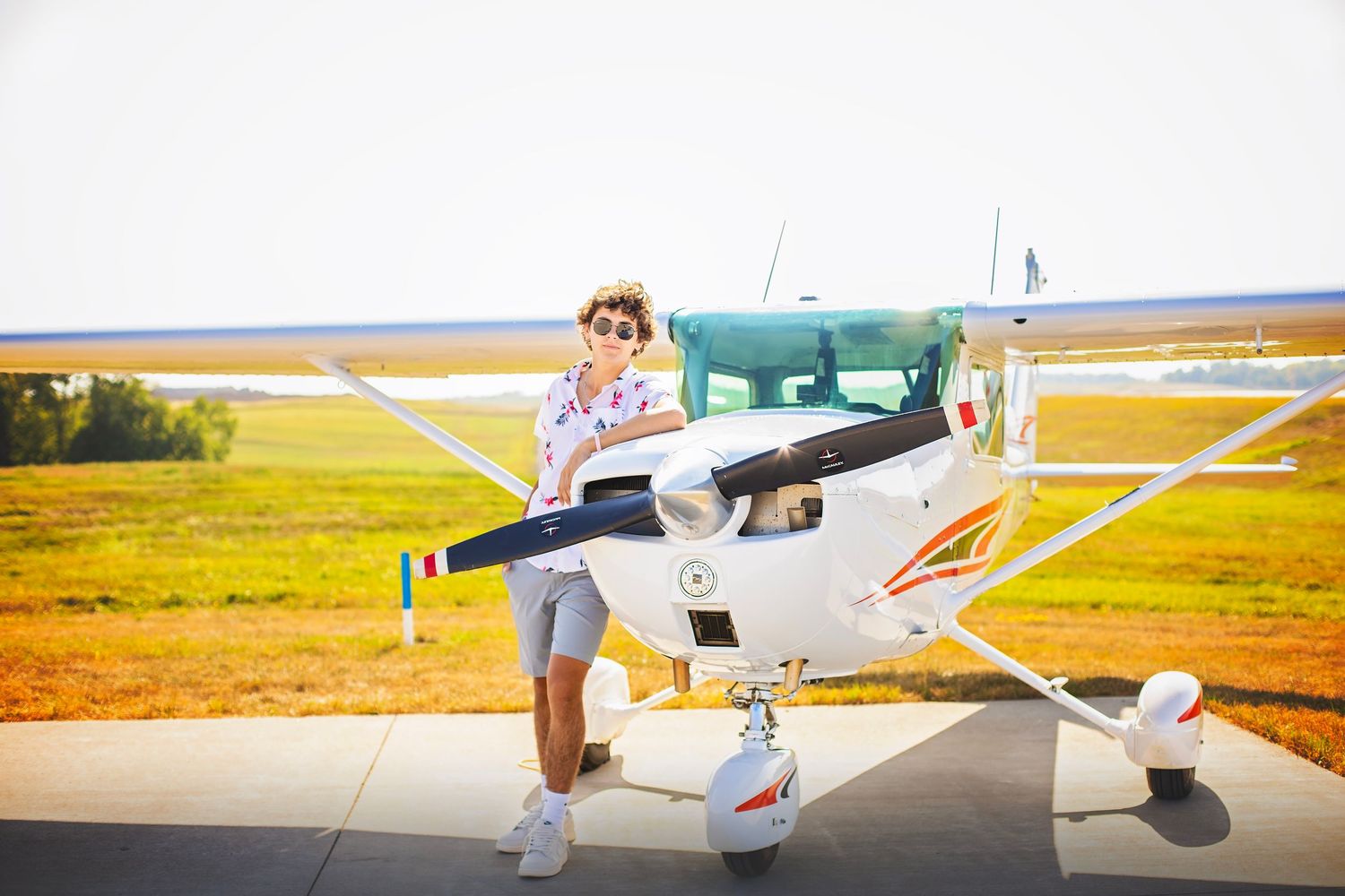 Teen boy in floral shirt stands beside a white propeller plane on a sunny airfield.