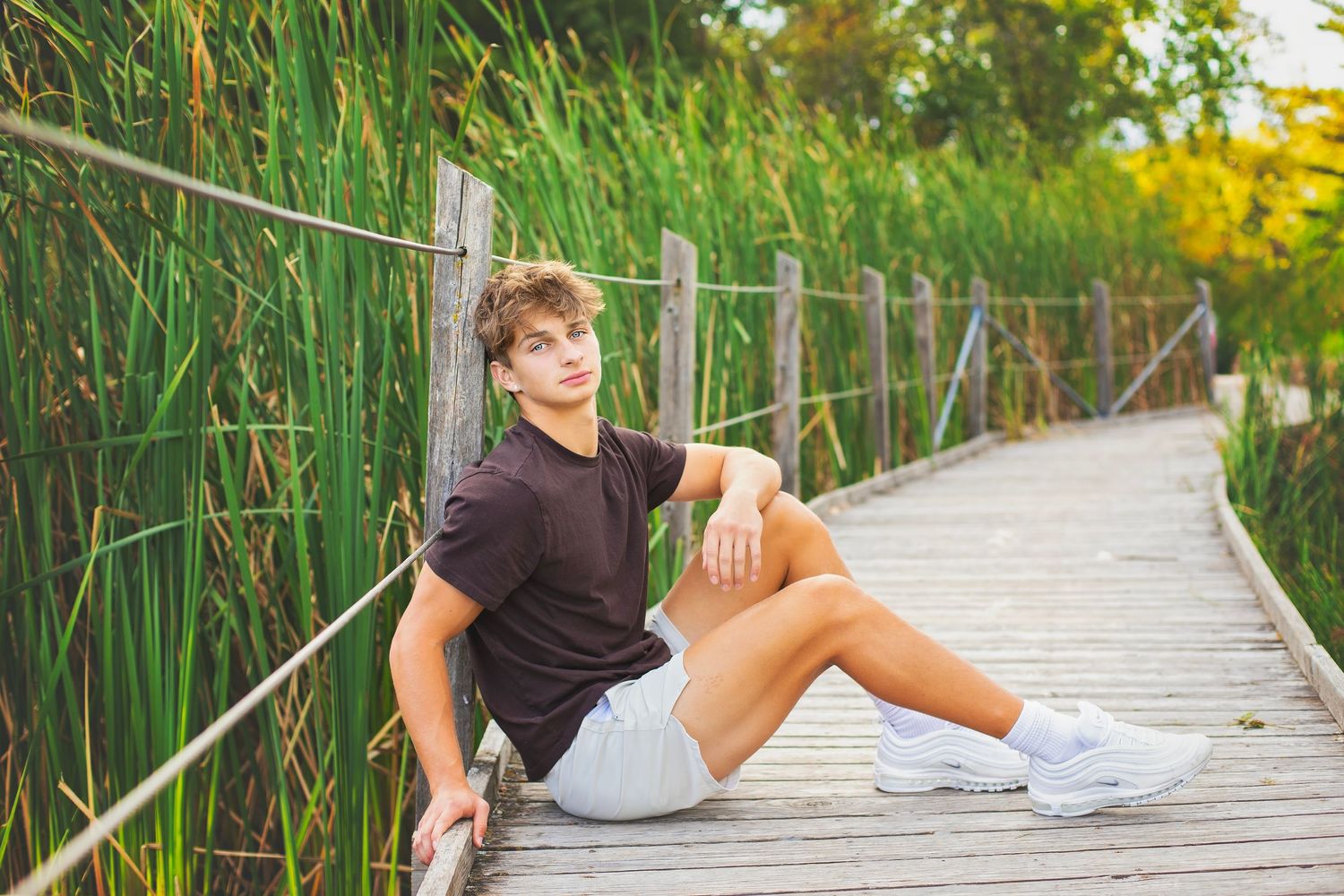 Teen boy in dark t-shirt and gray shorts sits on a wooden boardwalk surrounded by tall green reeds.