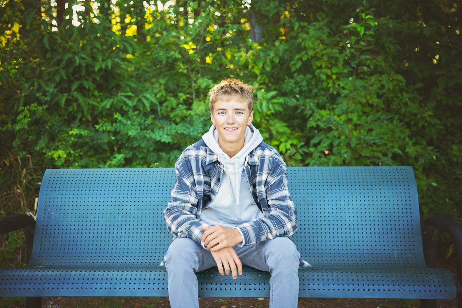 Smiling teen boy in plaid hoodie sitting on blue bench with green trees in background.