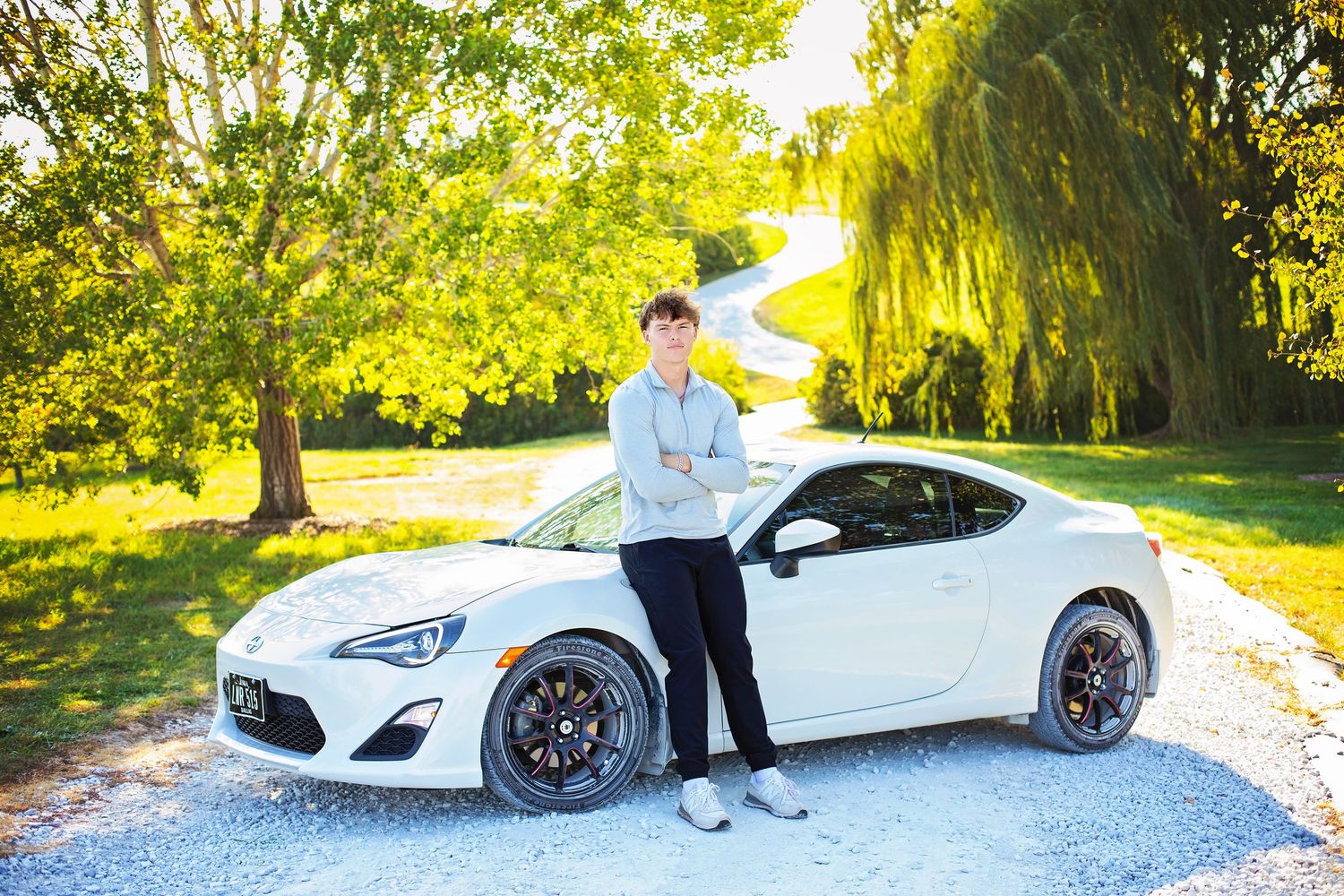 Teen boy poses beside a white Toyota 86 sports car on a tree-lined gravel driveway.