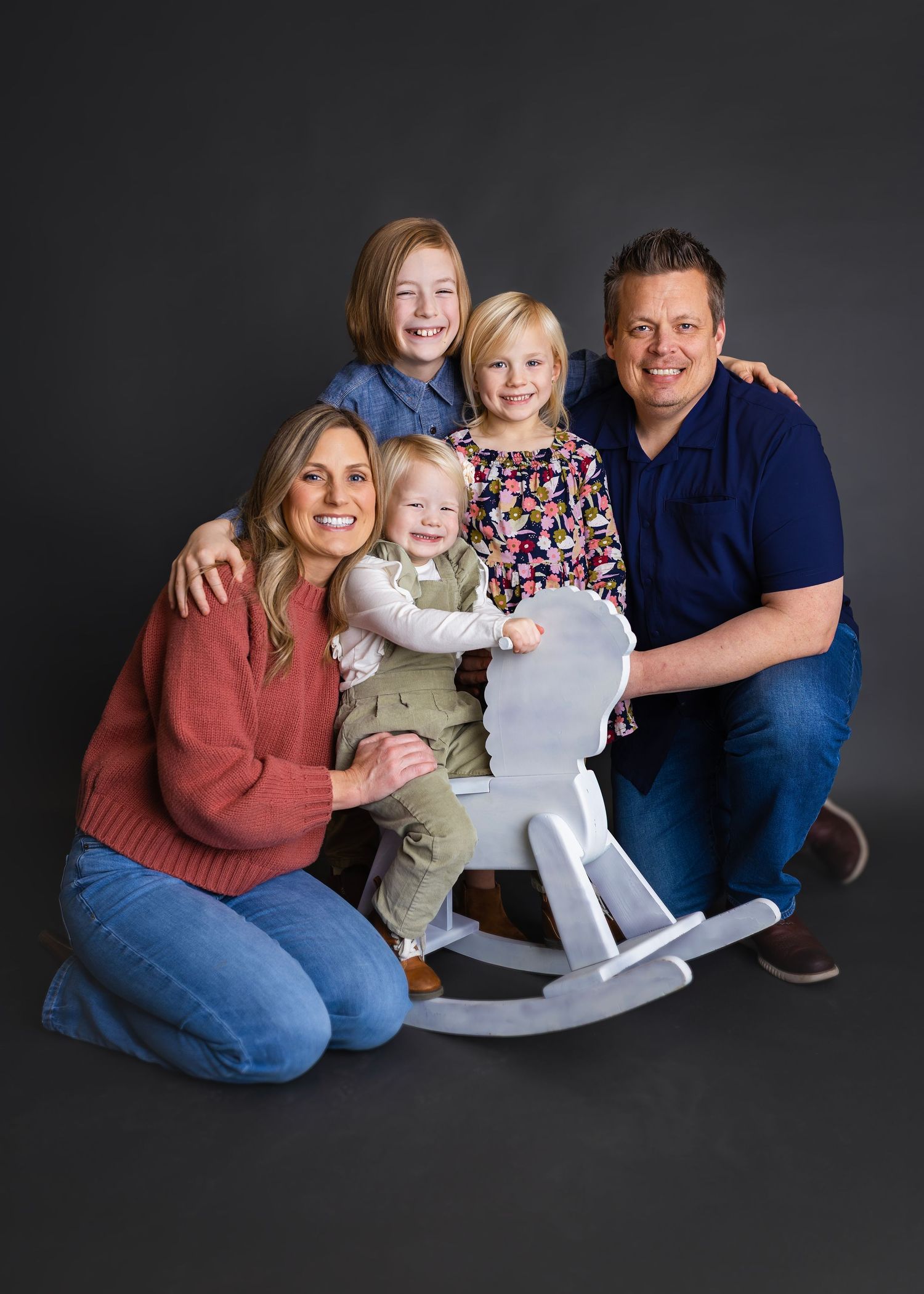 Smiling family of five posing with a white rocking horse toy against a dark gray background.