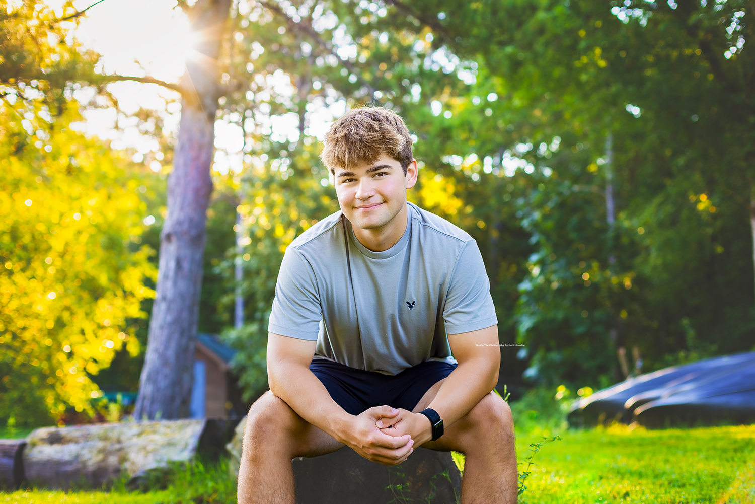 Young man in gray t-shirt sitting outdoors on rock, smiling, surrounded by lush green trees in golden sunlight.