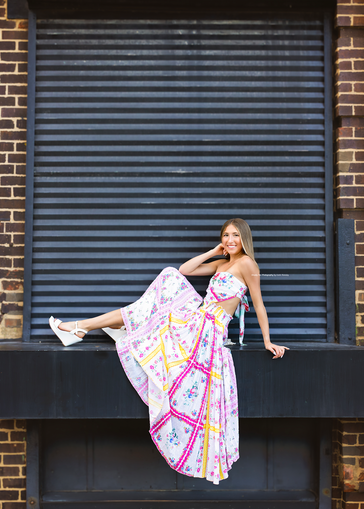 Woman in floral maxi dress poses on urban ledge with cocktail glass against brick wall backdrop.