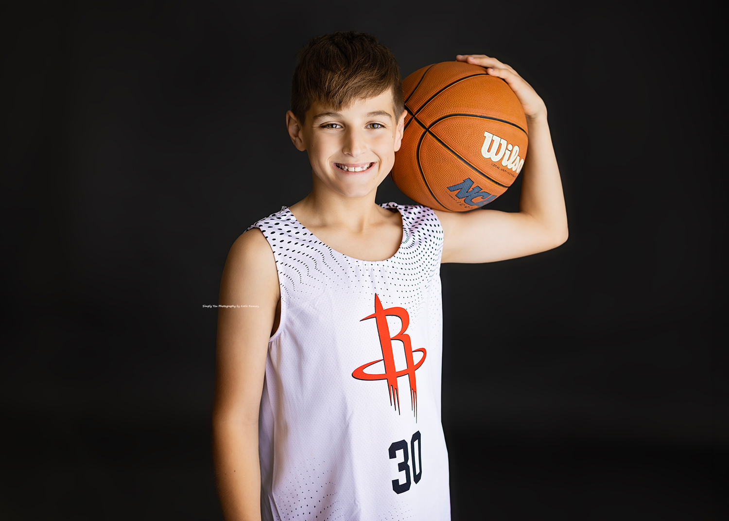 Smiling boy in Houston Rockets jersey #30 holding a Wilson basketball on his shoulder against dark background.