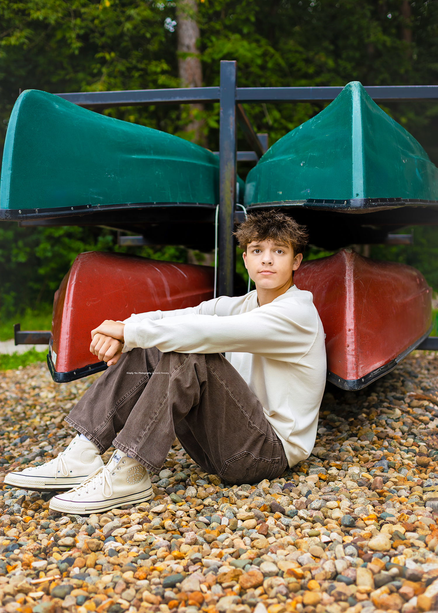 Teen boy in white hoodie and brown pants sits on gravel in front of stacked green and red canoes outdoors.