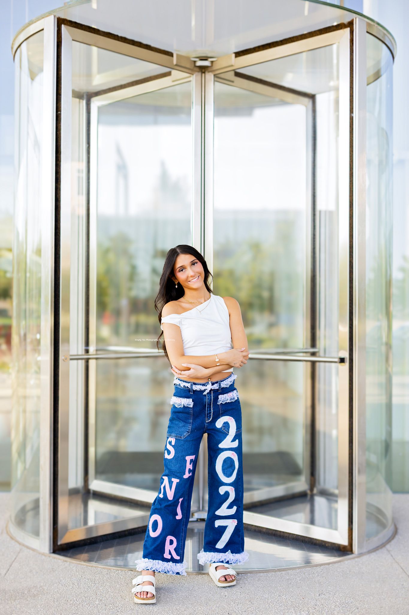 Teen girl in white crop top and blue Senior 2027 wide-leg jeans poses in front of glass revolving door.