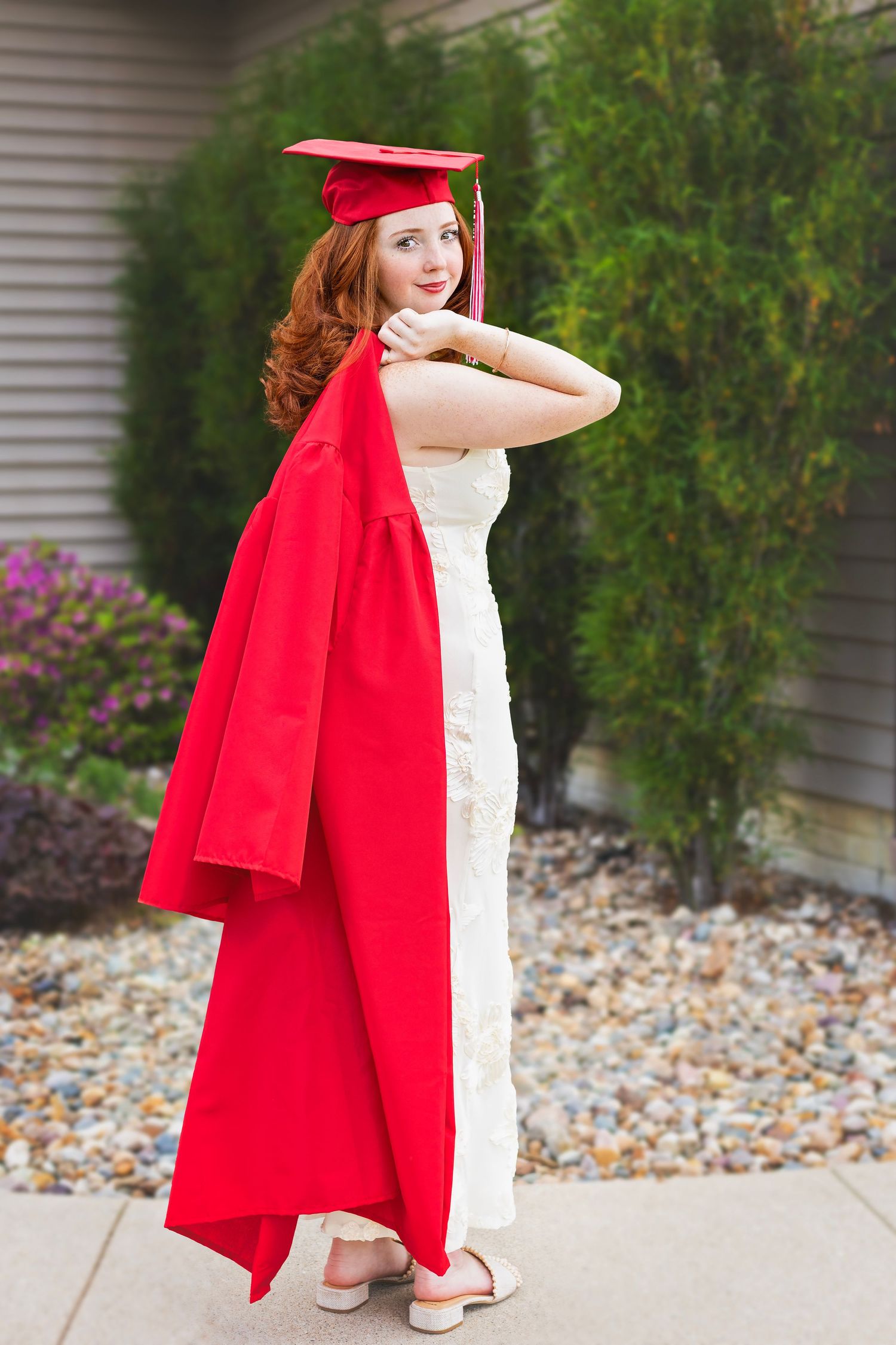 Young woman in red graduation cap and gown over white dress, posing outdoors near green shrubs.