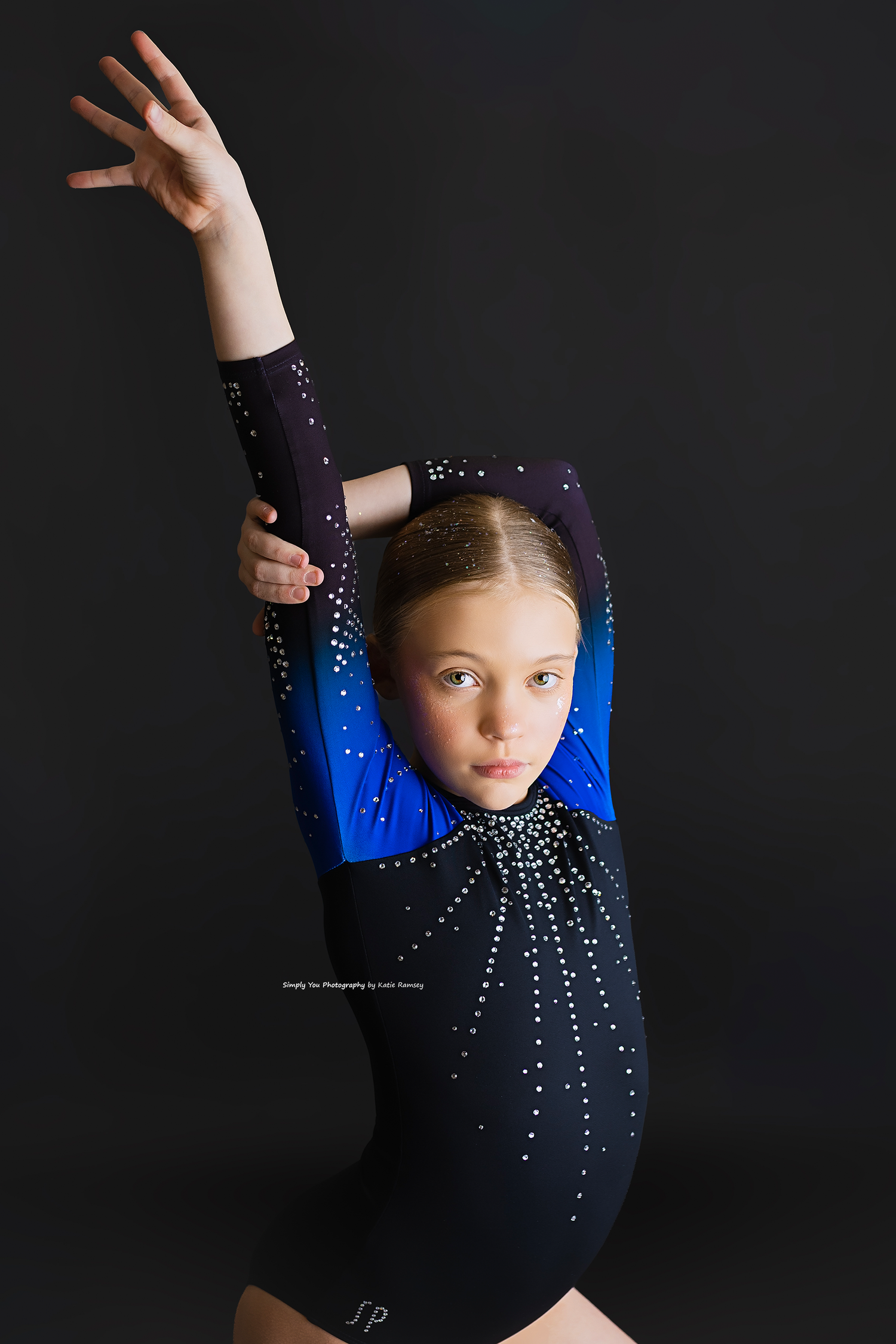 Young gymnast in a black and blue rhinestone leotard poses gracefully with arm raised against a dark background.