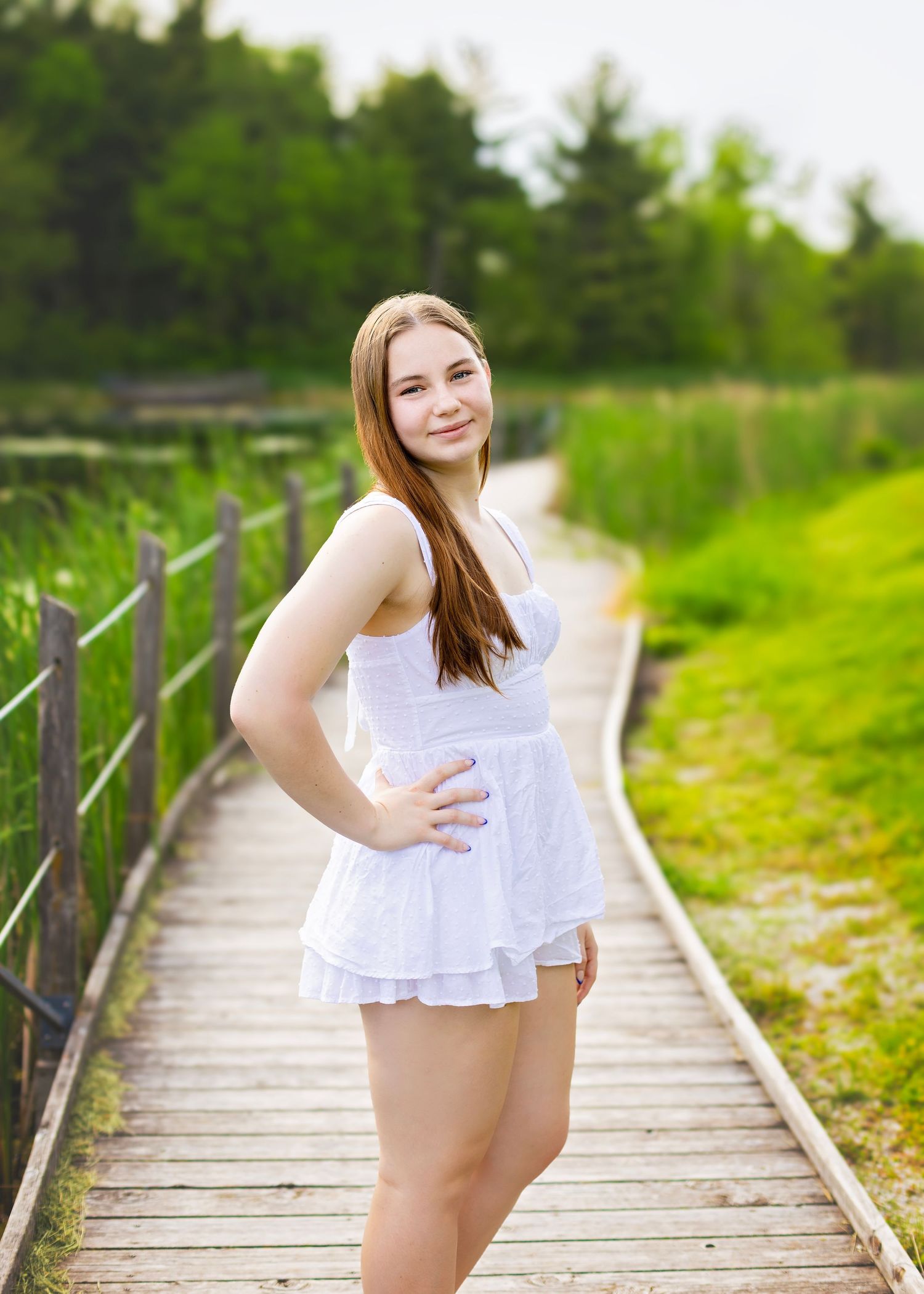 Young woman in white dress standing on wooden boardwalk with green trees and pond in background.