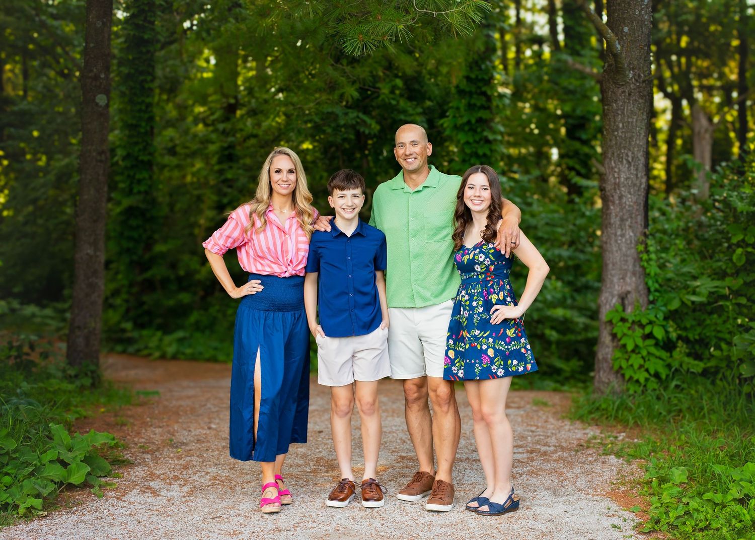 Family of four posing together on gravel path surrounded by tall green trees outdoors.