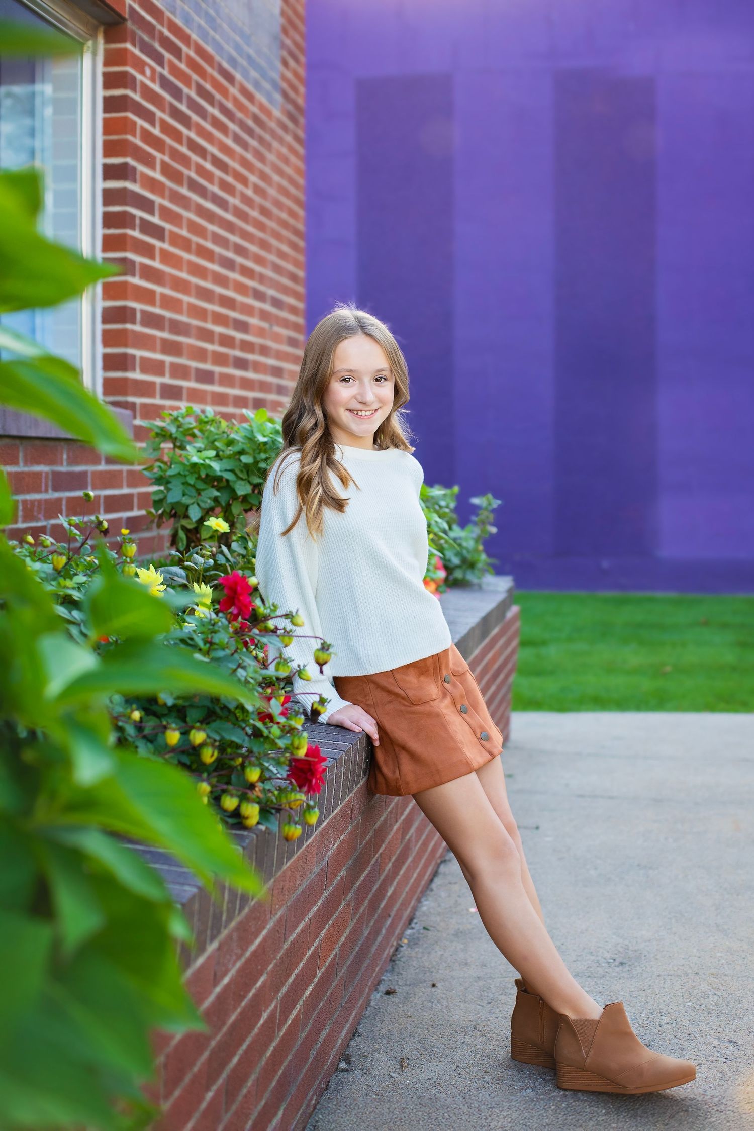 A young smiling girl in a white top and brown skirt leans against a brick planter with flowers.