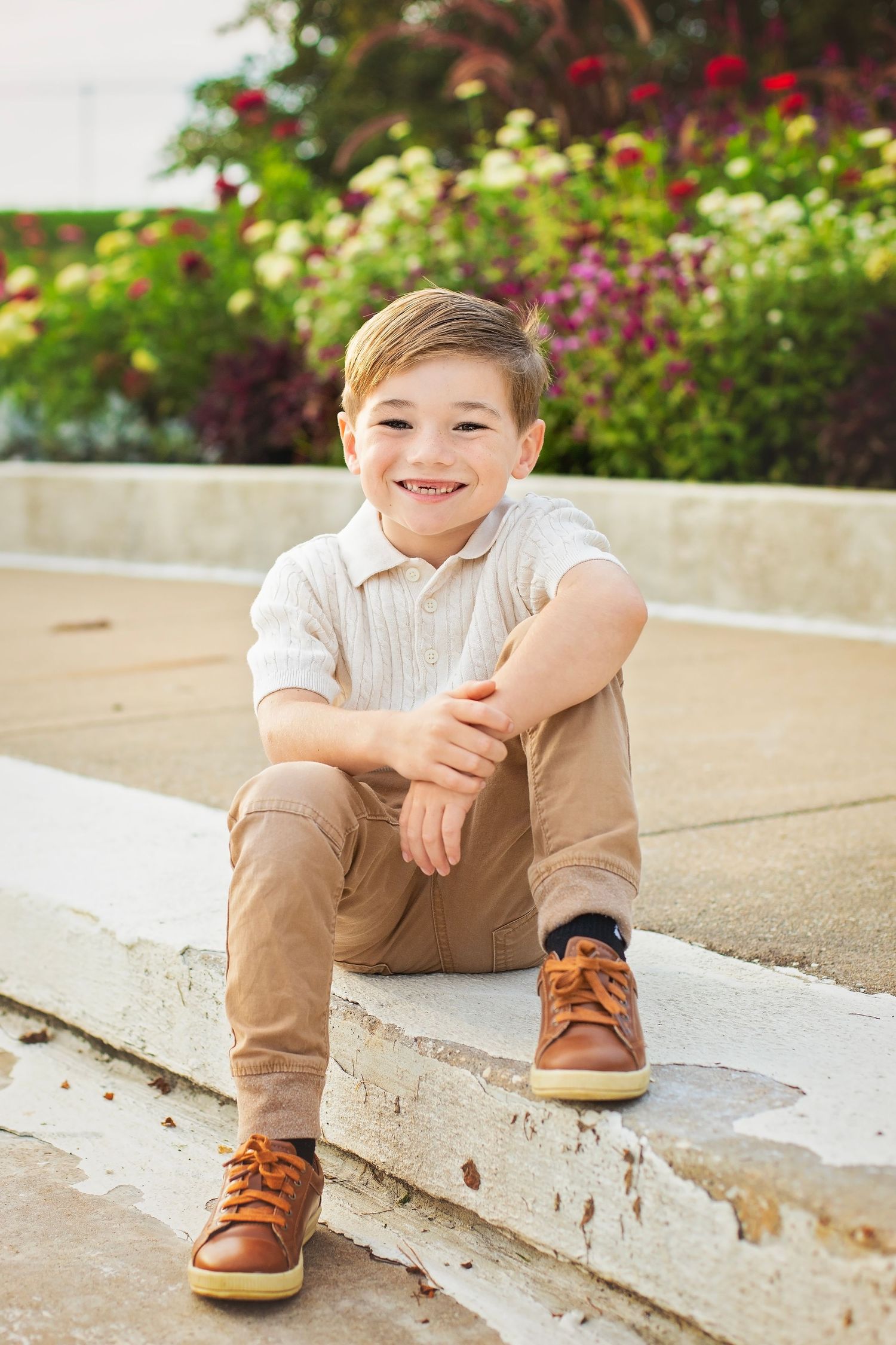 Adorable young boy smiling while sitting on stone steps with colorful flowers in the background.