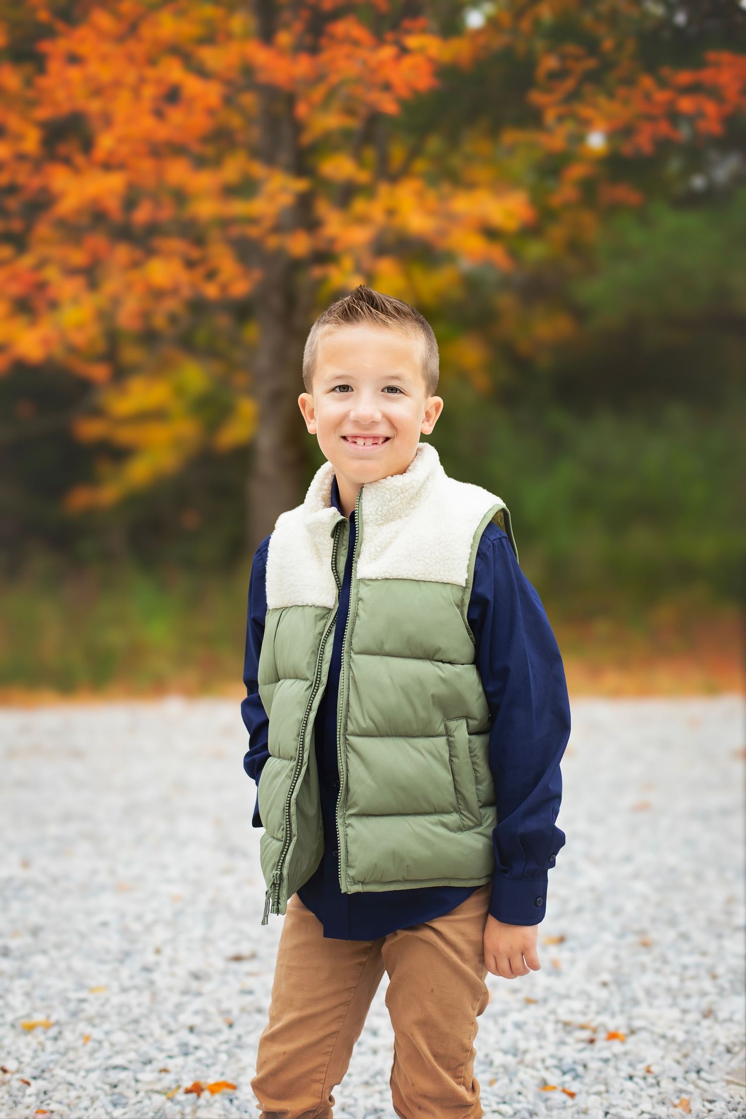 A young boy in a green vest and navy sweater smiles outdoors with colorful fall foliage behind him.