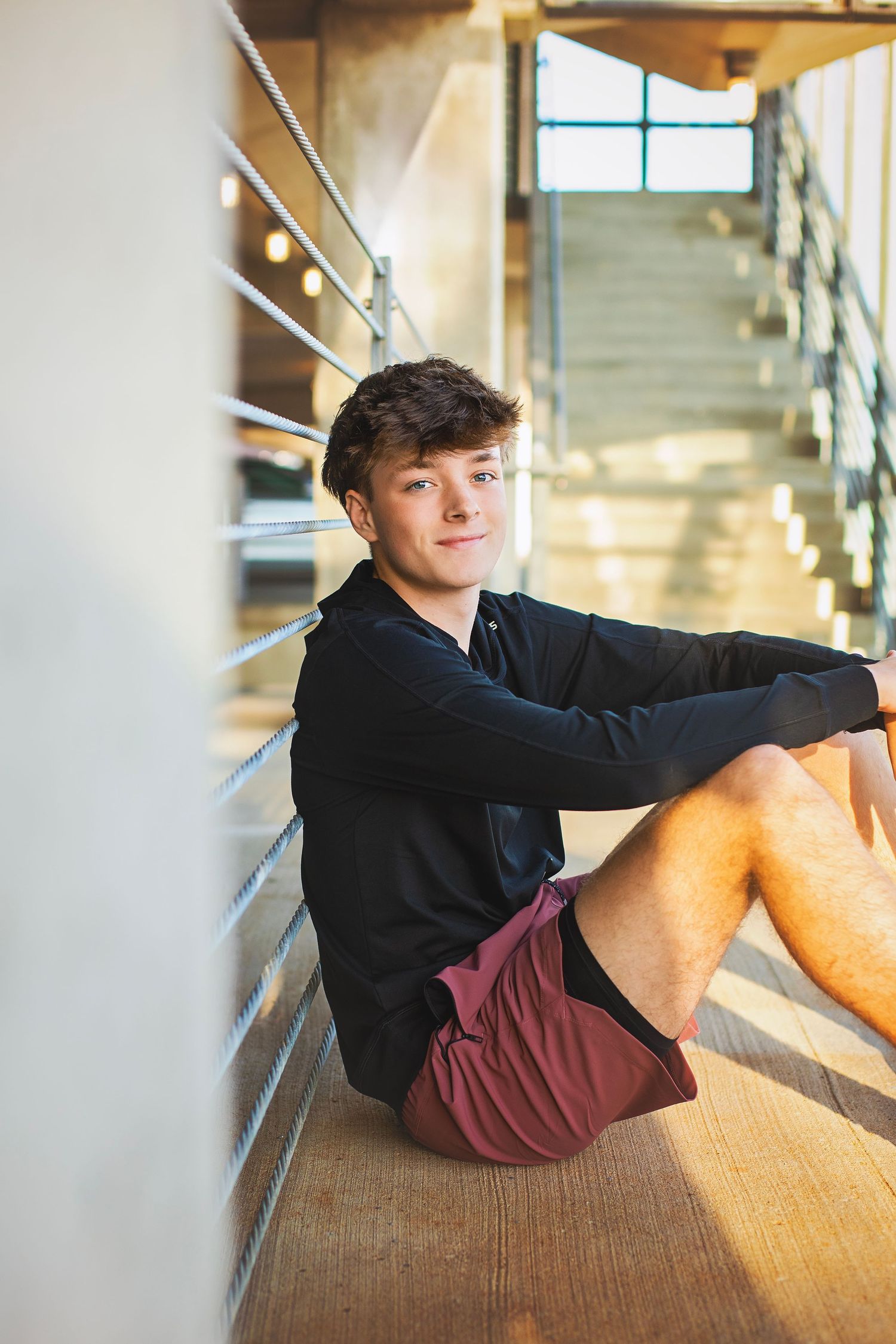 Teen boy in black hoodie and maroon shorts sitting on floor leaning against wall near staircase.