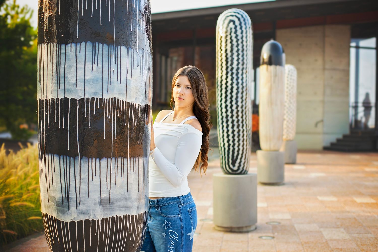 Young woman in white off-shoulder top leans against decorative column near large cactus sculptures outdoors.