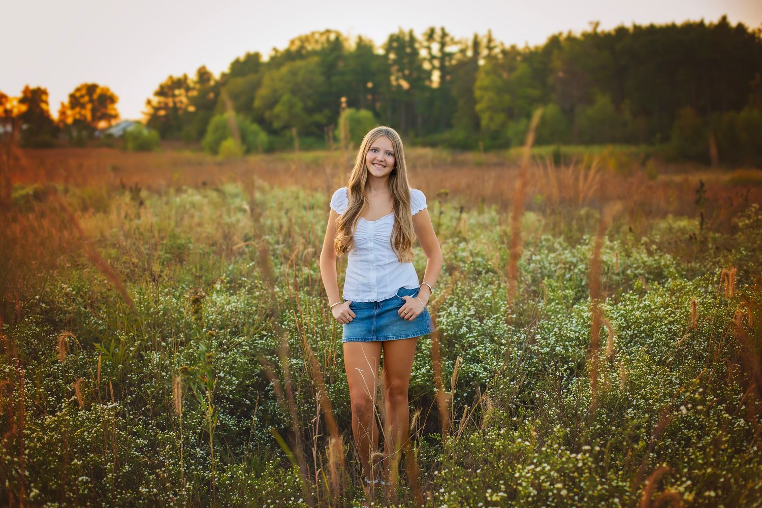 Smiling blonde teen in white tank and denim shorts stands in wildflower meadow at sunset.