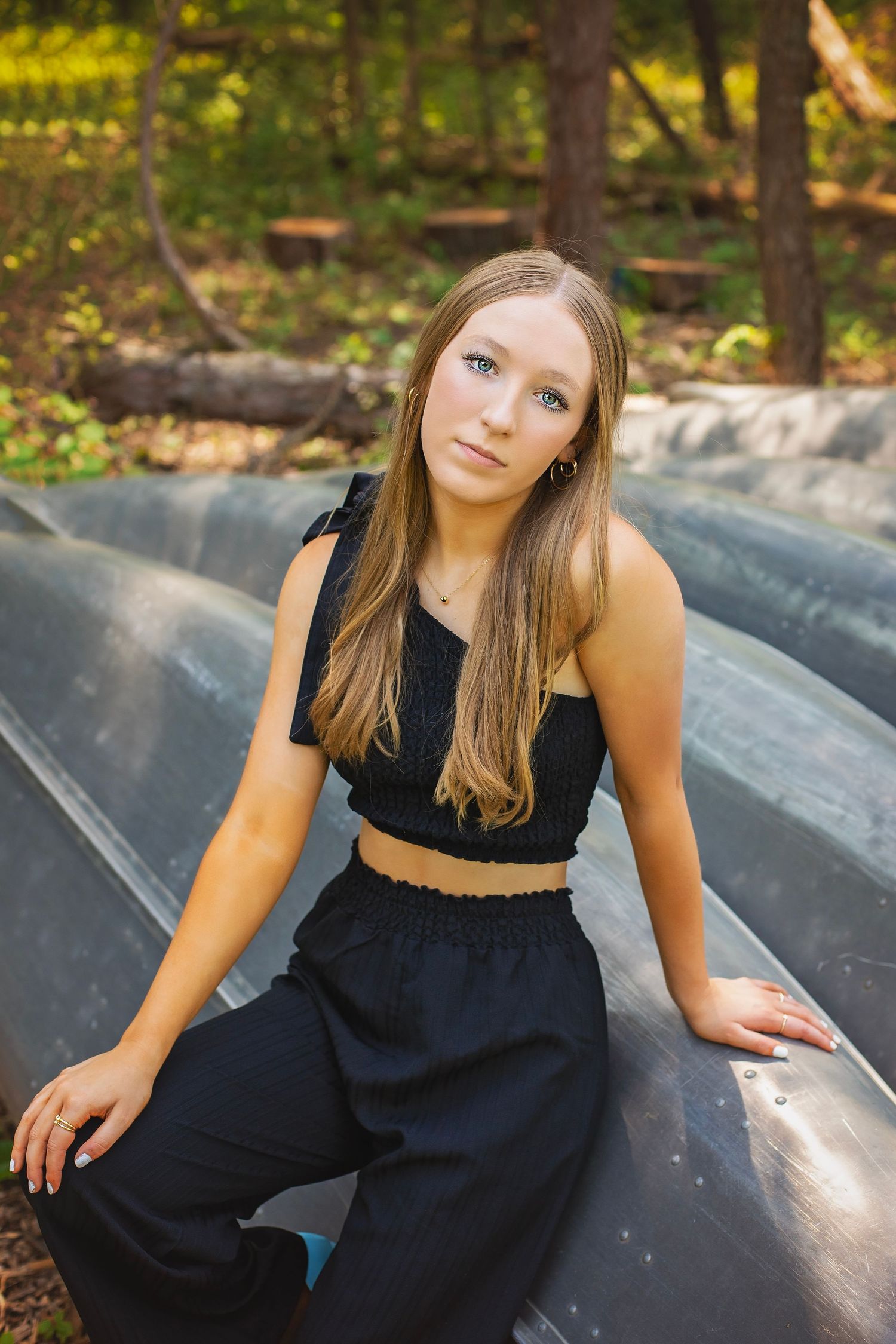 Teen girl in black crop top and wide-leg pants poses on metal slides in a wooded outdoor setting.