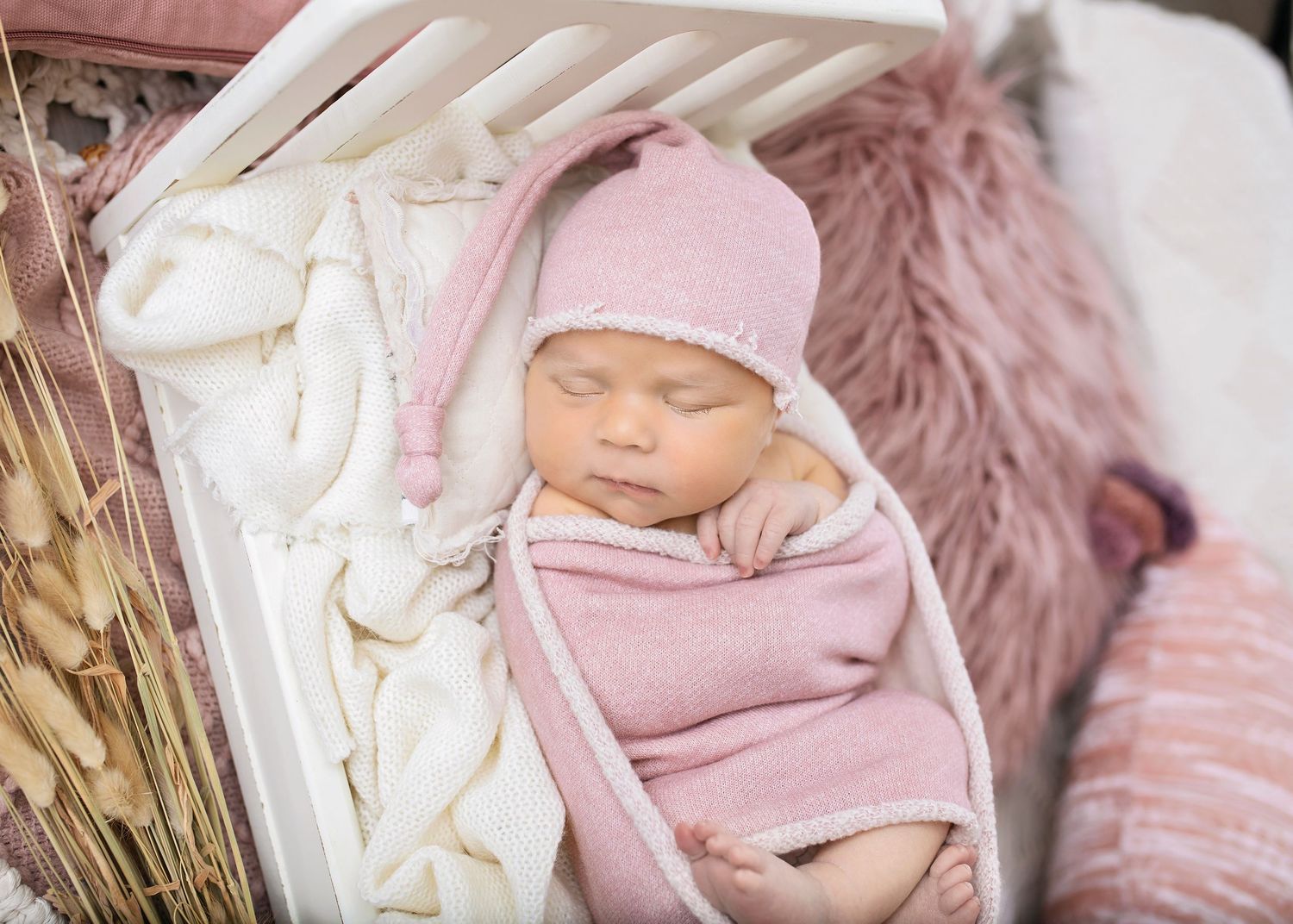 Sleeping newborn baby girl swaddled in pink wrap wearing knit hat resting in white mini crib.