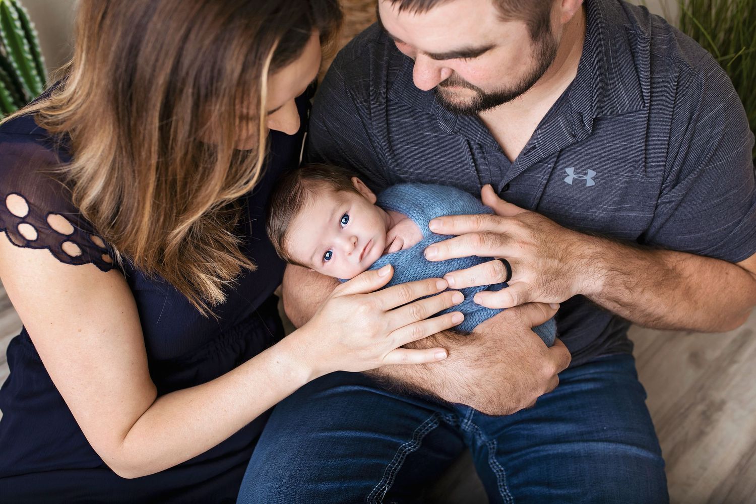 Parents lovingly cradling their wide-eyed newborn baby boy wrapped in blue swaddle cloth.