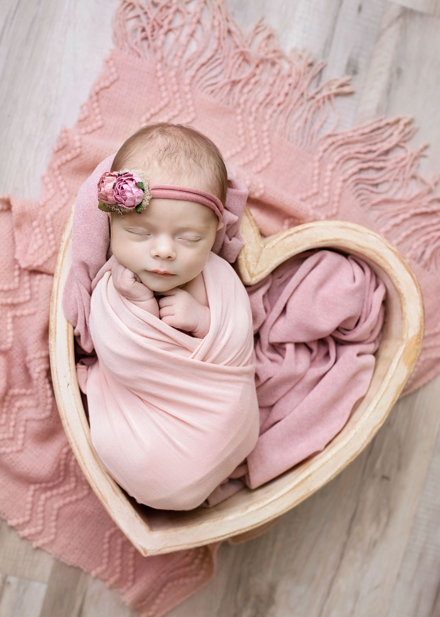 Newborn baby girl wrapped in pink swaddle with floral headband sleeping in heart-shaped wooden bowl.