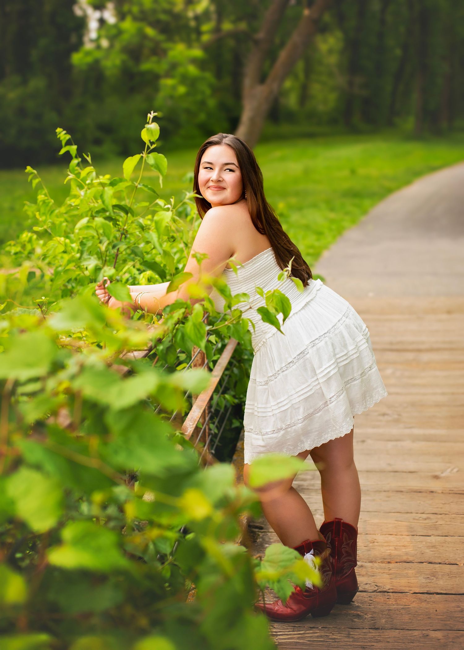 Smiling young woman in white dress and cowboy boots poses on a wooden path in lush greenery.