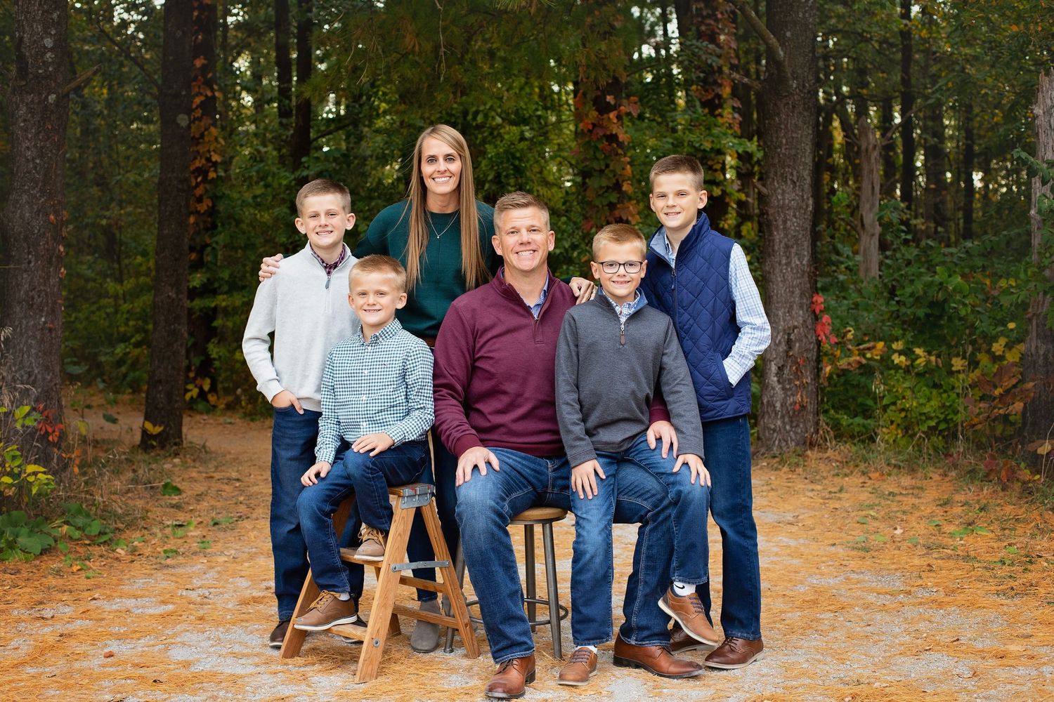 Family of six posing on wooden stools in a colorful autumn pine forest.