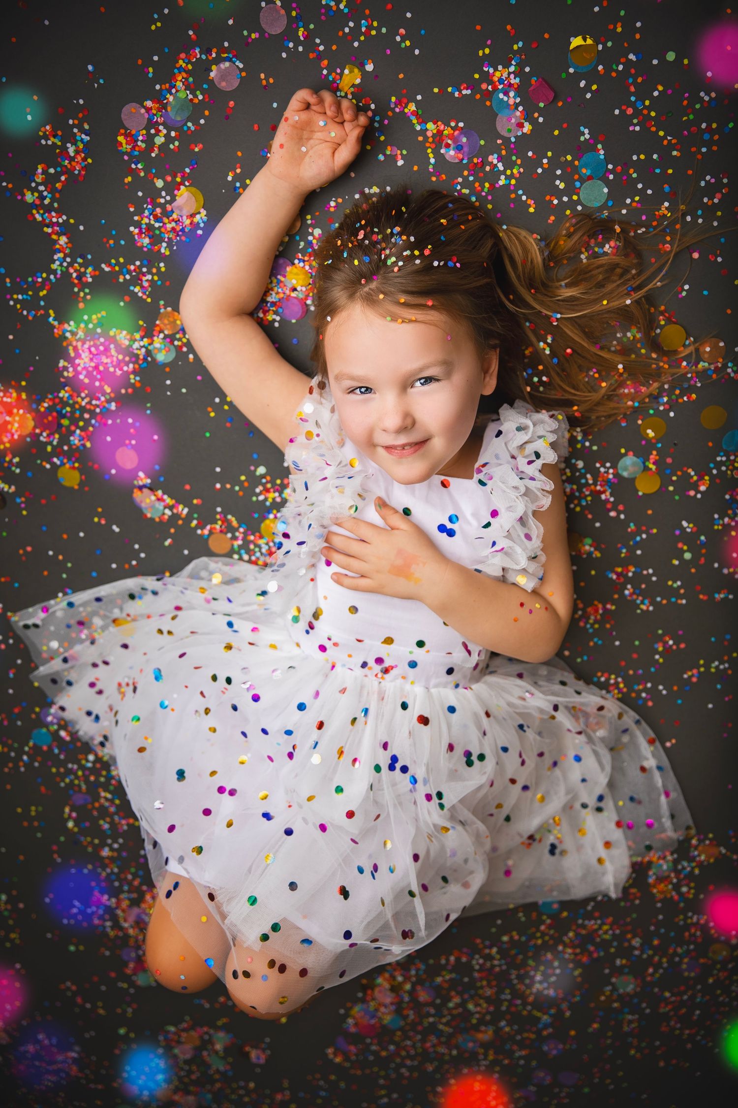 A joyful young girl in a white dress lies on the floor dancing among colorful confetti and bokeh light effects.