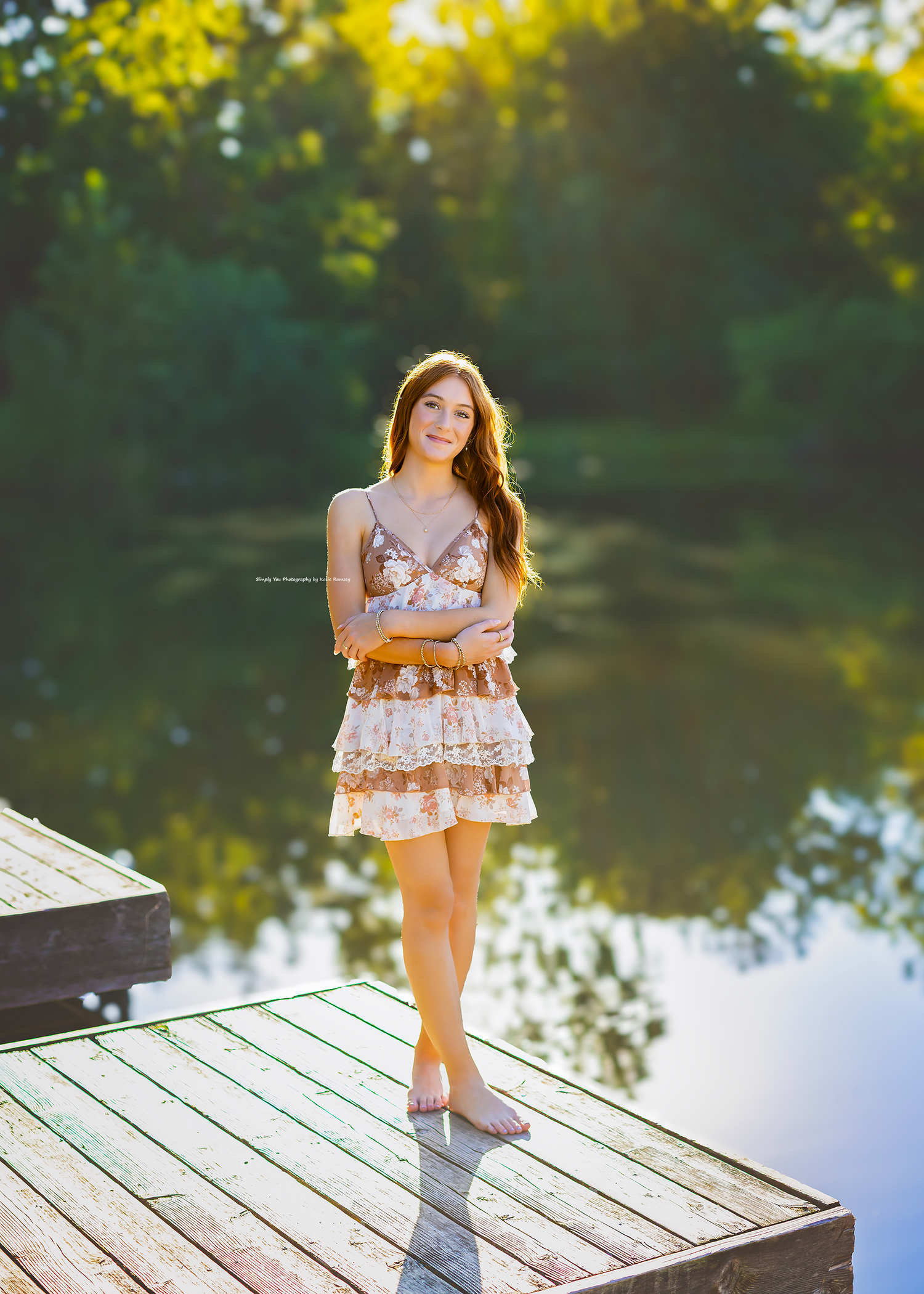 Young woman in floral dress standing on wooden dock by serene reflective lake surrounded by lush green trees in golden light.
