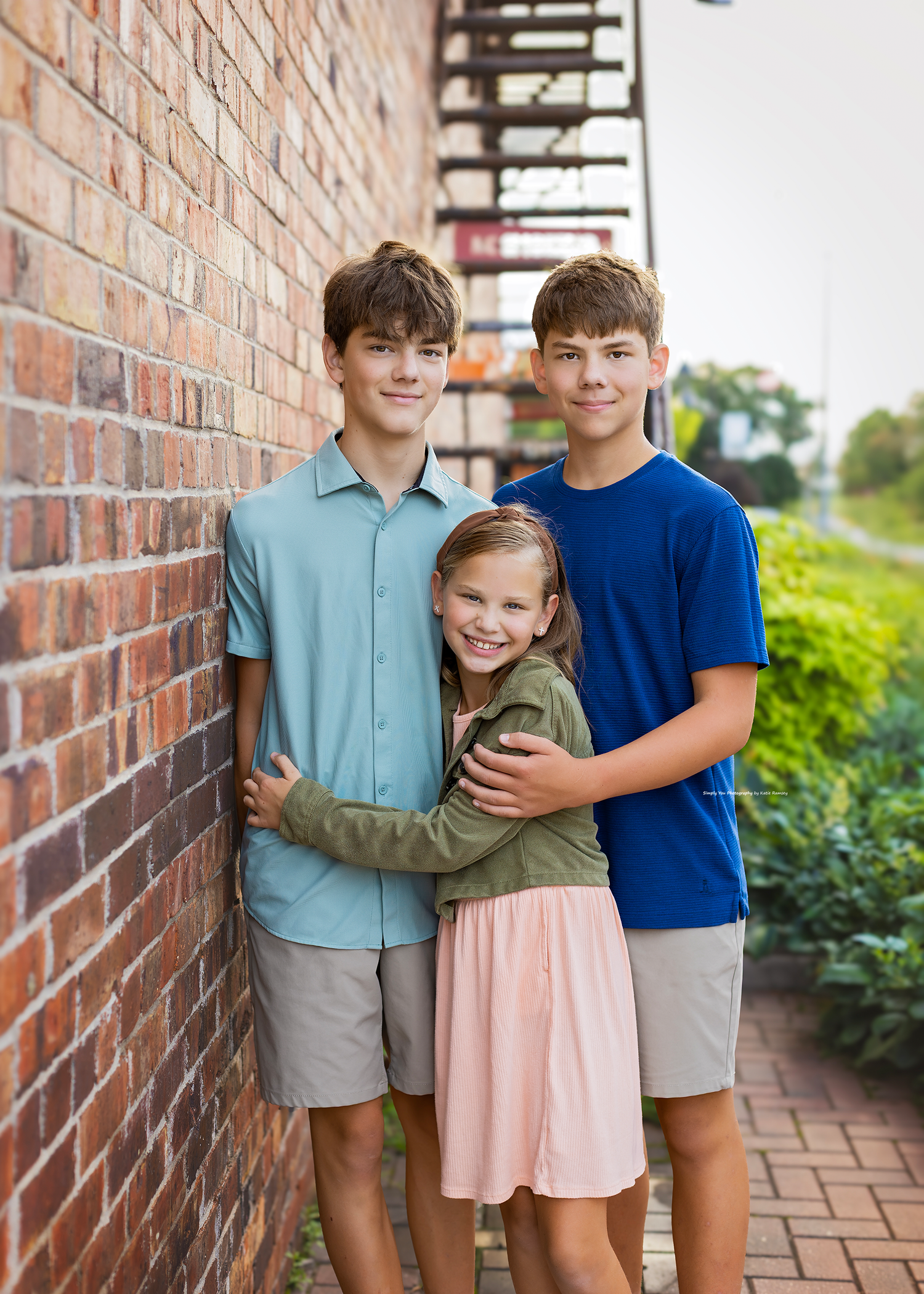 Three siblings pose together against a brick wall outdoors, two older boys flanking a smiling young girl in a pink dress.