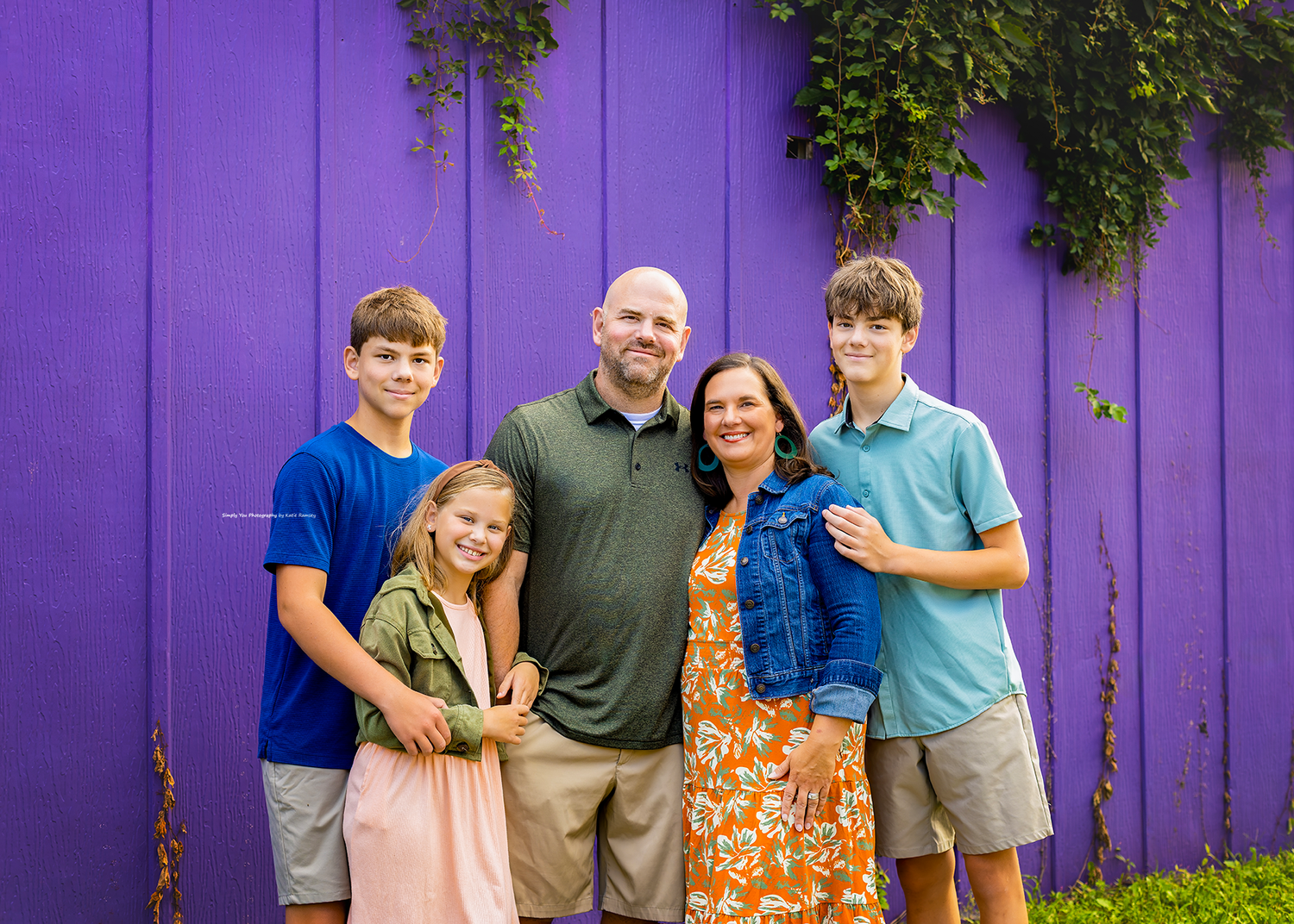 Family of five smiling together in front of a vibrant purple wooden wall with green ivy.
