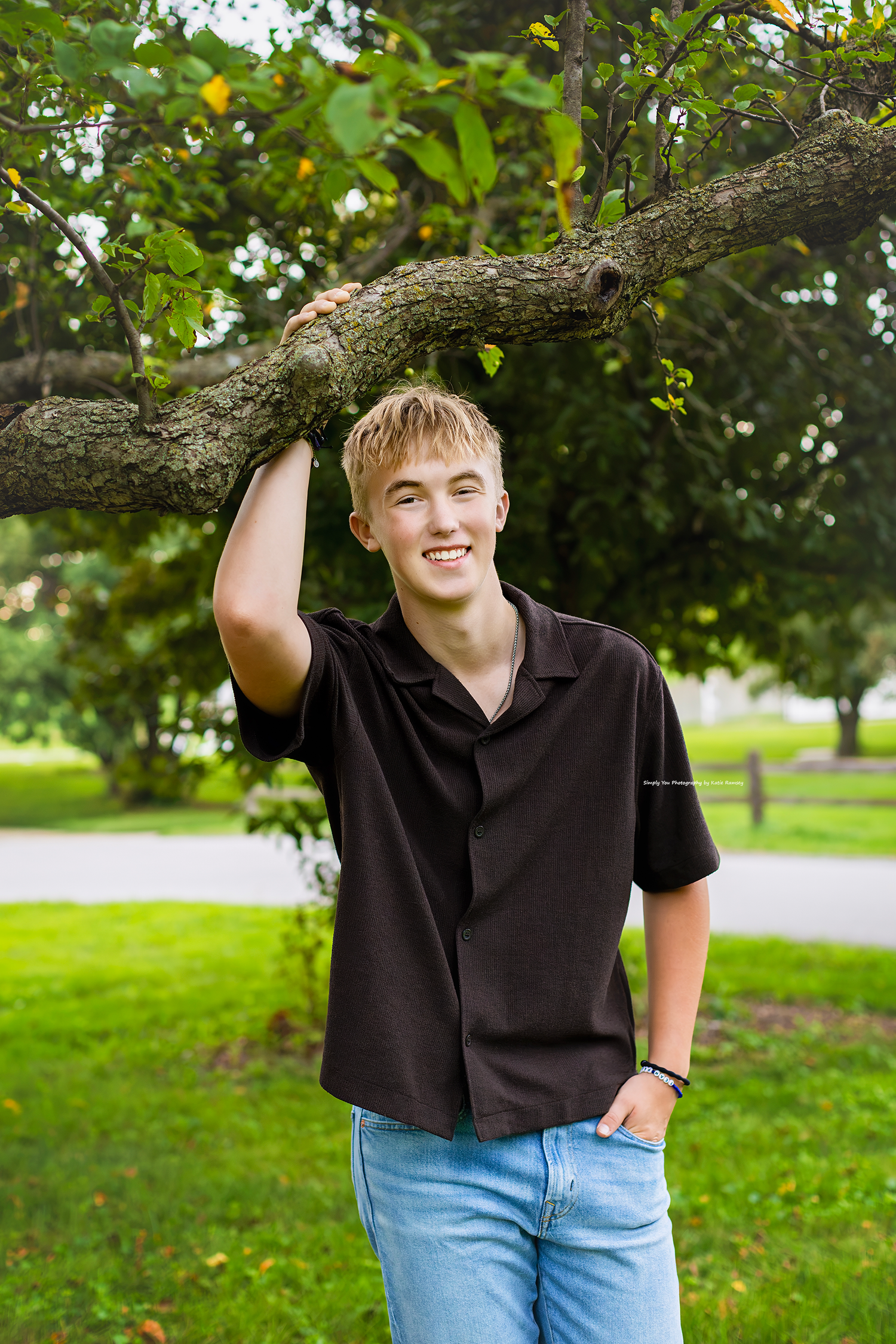 Smiling teen boy in black shirt and jeans leaning against a tree branch in a lush green park setting.